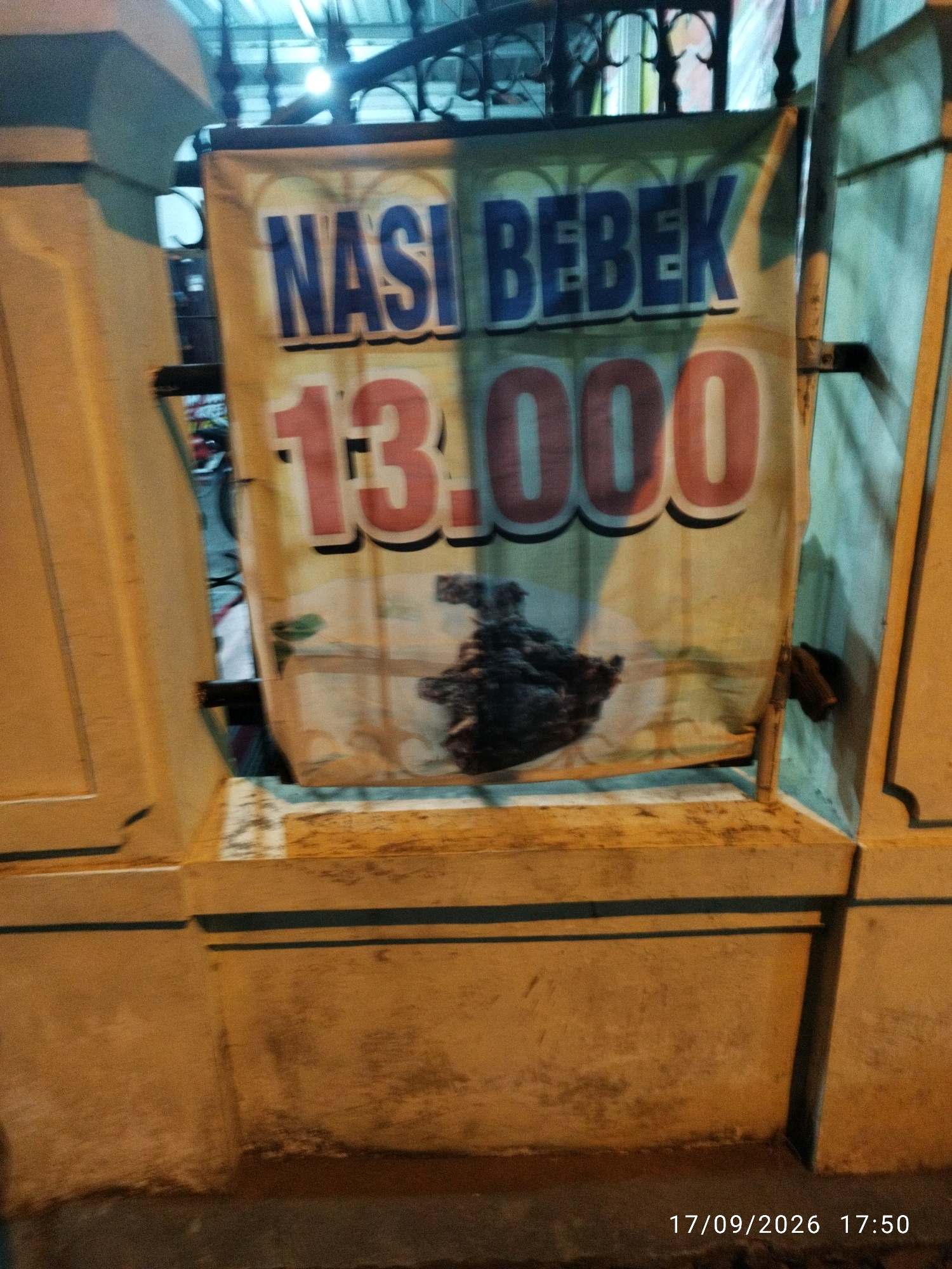 A close-up, slightly blurry, eye-level, nighttime exterior shot shows a banner advertising "NASI BEBEK" (duck rice) for 13,000. The banner is yellow and features the text in large blue letters for "NASI BEBEK" and red numbers for the price. Below the text, there is an image of what appears to be cooked duck meat. The banner is hanging from a metal support structure, and it is partially obscured by a yellow concrete wall in the foreground and a blue-green wall in the background. The scene is lit by artificial light, creating shadows and highlights. The timestamp in the bottom right corner indicates the date as 17/09/2026 and the time as 17:50. While the specific location within Mojokerto, Indonesia is not explicitly stated, the banner suggests a food stall or restaurant selling this particular dish.