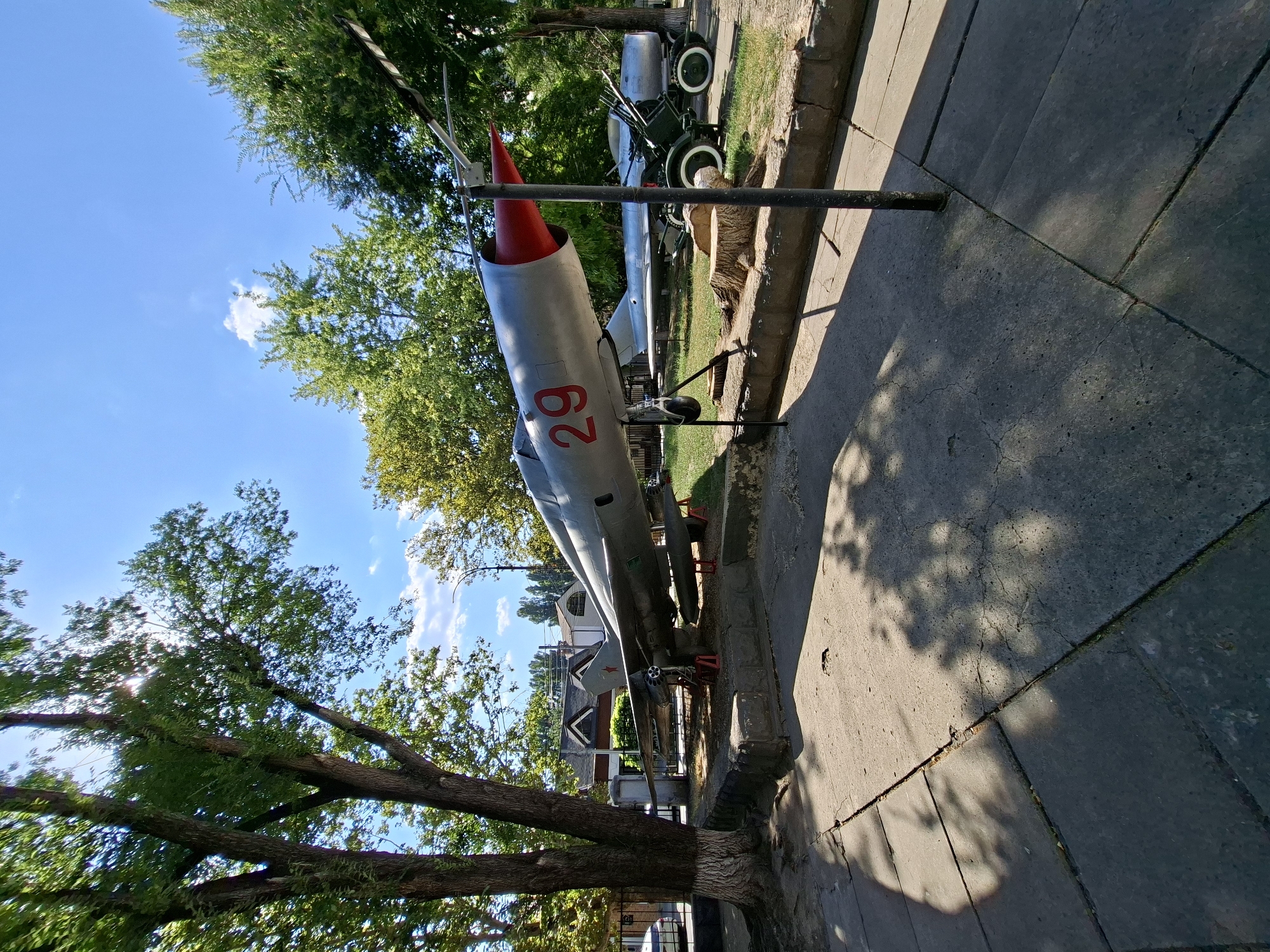 A jet fighter aircraft, painted silver with a red nose cone and marked with the number 29 in red, is displayed outdoors on a sunny day. The aircraft is positioned on display stands in a park-like setting with mature trees and a clear blue sky overhead. In the background, a military cannon with wheels is also visible, along with some buildings and a fence. The foreground shows a paved walkway and some tree stumps. This image was taken in Chișinău, Moldova, at the Museum of Military History.