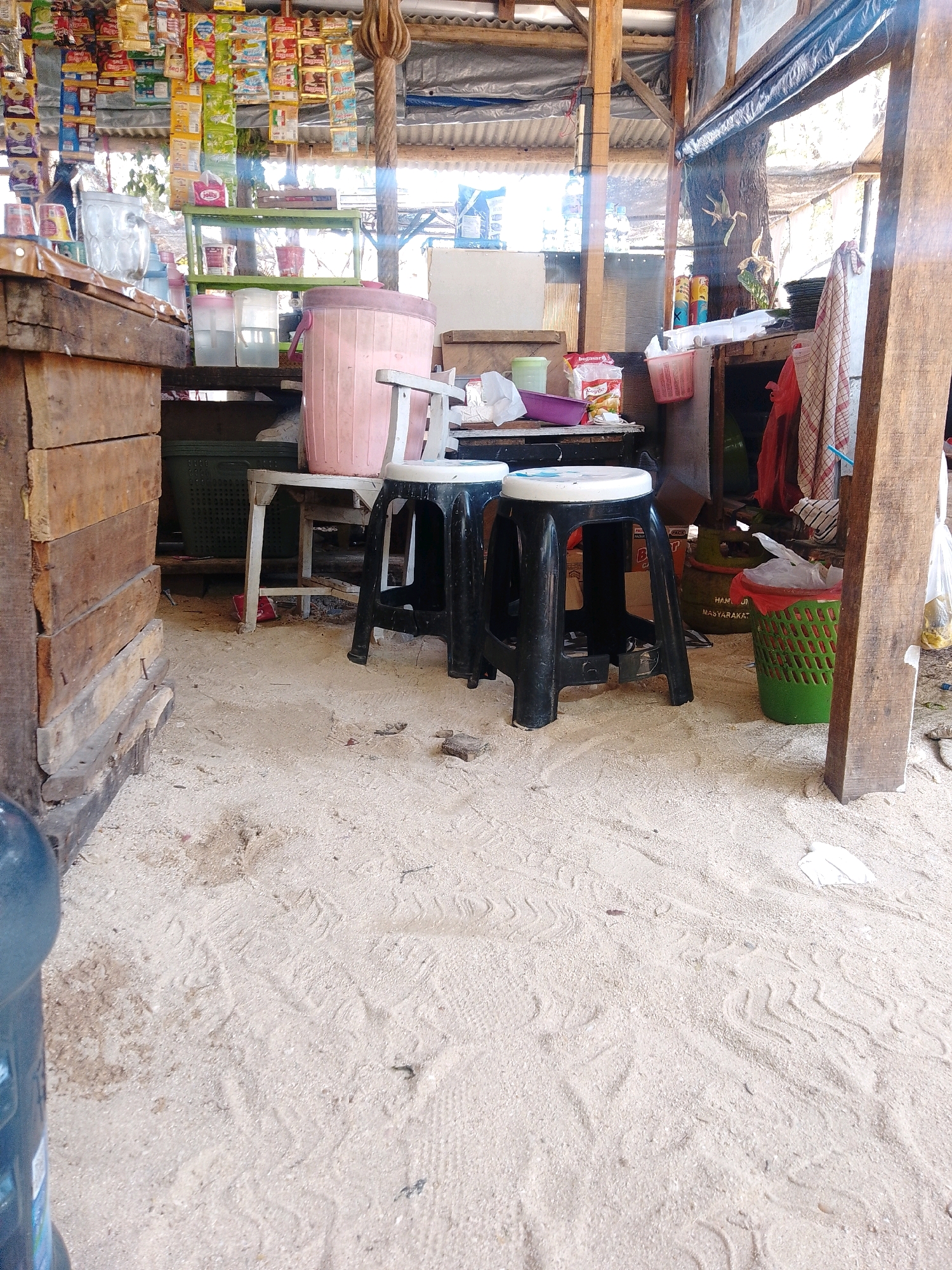 The image depicts the interior of a rustic, open-air roadside stall or small shop, likely in Cikal, Indonesia, given the context. The floor is covered in a thick layer of light-colored sand. In the foreground, on the left, part of a large blue water jug is visible. The stall's construction is simple, with wooden planks forming walls and a slatted wooden roof. Shelves are stocked with various packaged goods, including snacks and drinks.
Dominating the center of the scene are two black plastic stools with white tops, placed in front of a counter. Behind the stools, a large pink plastic container is visible, alongside a white table and a green plastic basket. In the background, more shelves hold items, and a large green gas cylinder is present. The overall impression is of a simple, functional establishment catering to local needs, potentially a small warung or convenience store. The lighting suggests it is daytime, with sunlight filtering through the roof and open sides.