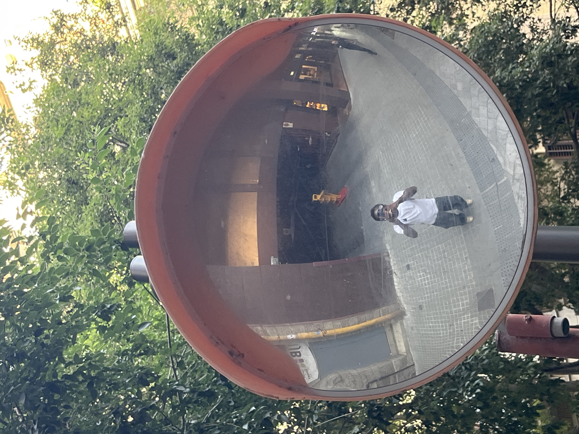 A wide-angle convex mirror, with an orange rim, is mounted on a pole in an urban environment, reflecting trees and buildings. The mirror is positioned at eye-level. In the reflection, a person wearing a white t-shirt and dark pants is visible, crouching and holding a smartphone to take a selfie. The background of the reflection shows a paved walkway, a building facade with dark wood panels and a yellow pole, and further in the distance, a bright yellow traffic cone on a red base. The overall scene appears to be in a pedestrian area or a street with limited vehicular traffic, possibly in Barcelona, Spain, given the architectural style and foliage. The lighting suggests it is daytime, with sunlight filtering through the trees.