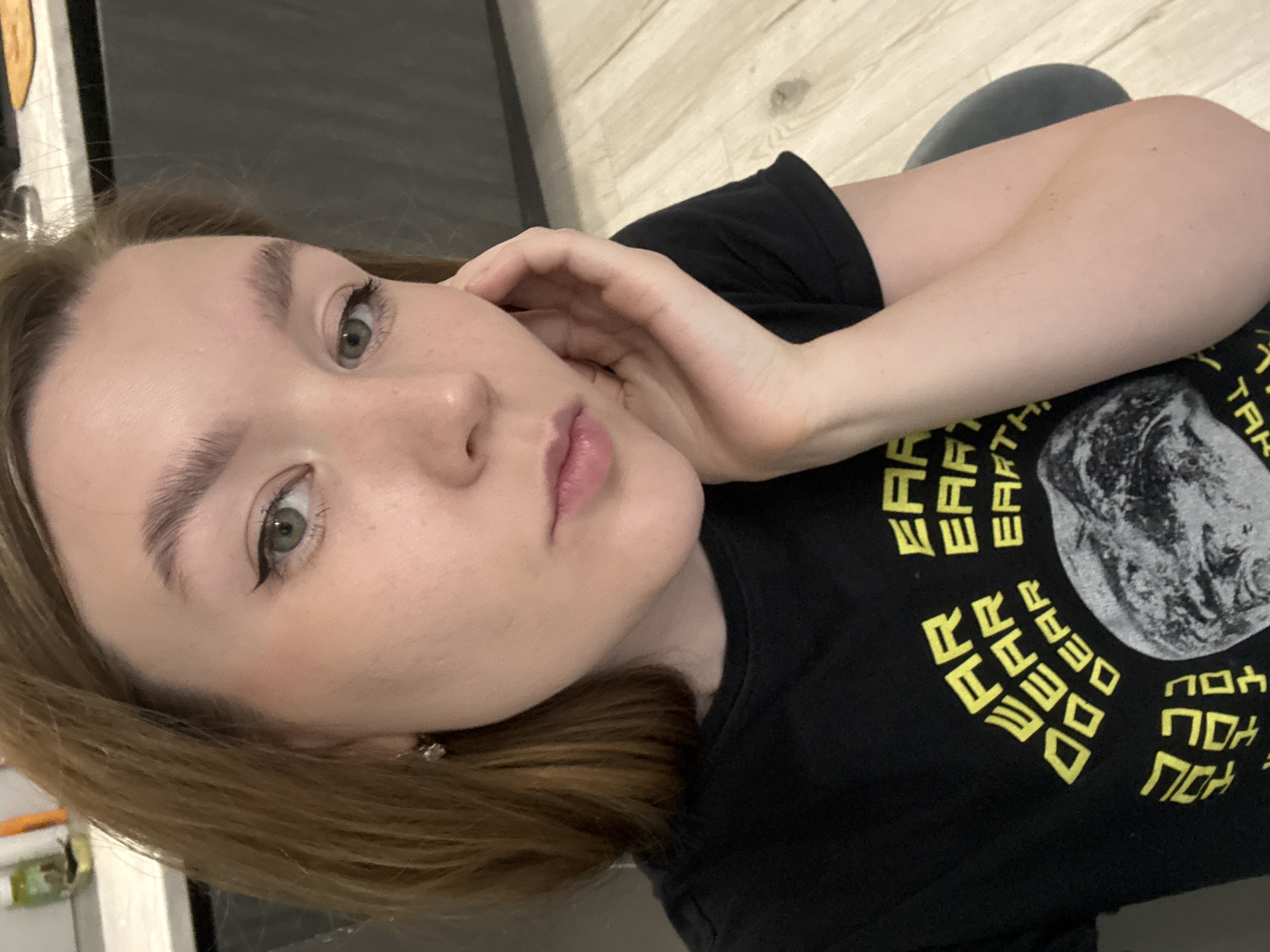 The image shows a woman with light skin, light brown hair, and green eyes. She is wearing eyeliner and a black t-shirt with yellow text surrounding a black-and-white image. Her right hand is touching her face near her cheek. The background includes a light-colored wooden floor and a dark surface, potentially a piece of furniture. There is no indication of Izhevsk, Russia, or any other specific location.
