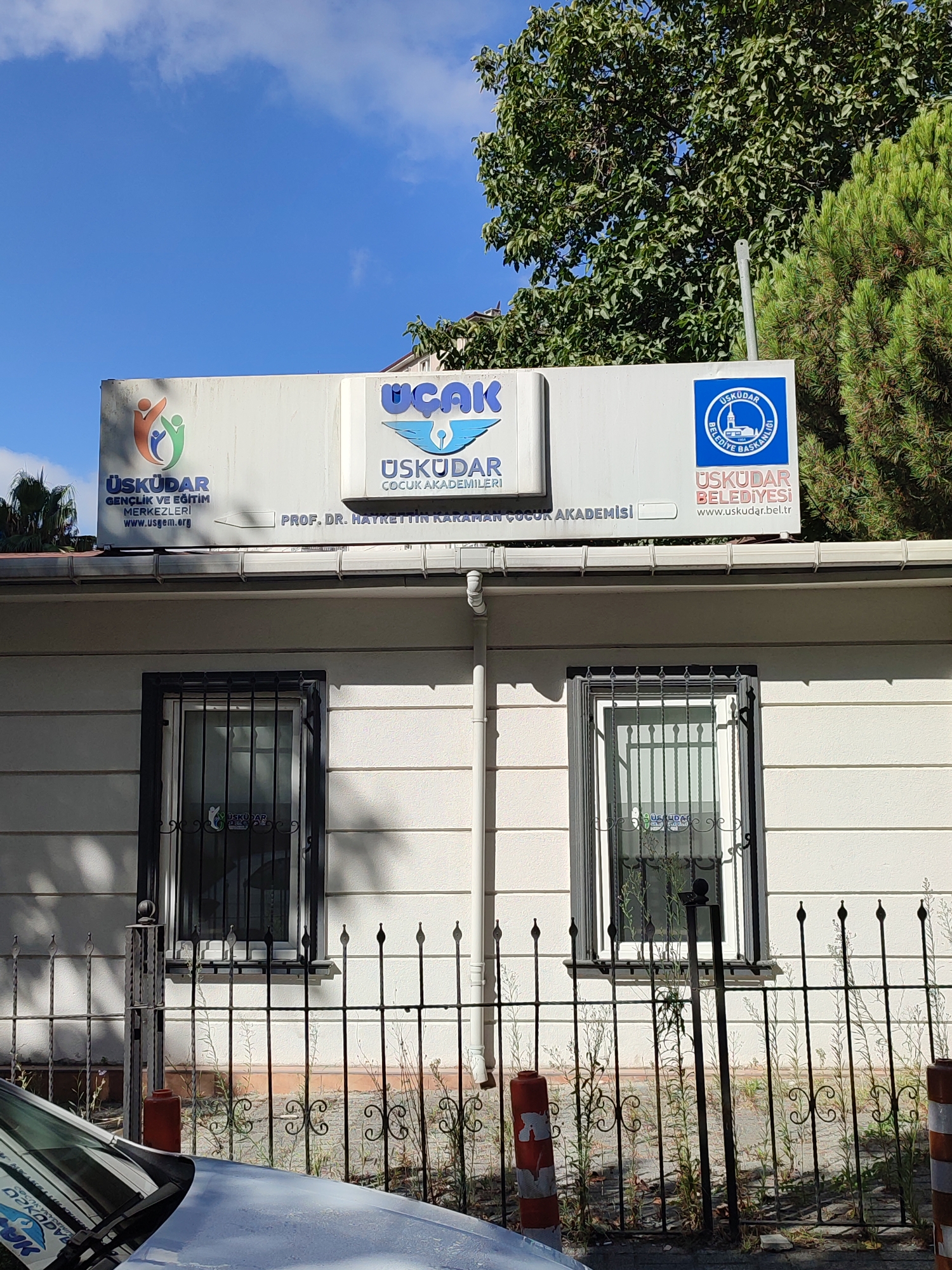 The image depicts the exterior of a building in Üsküdar, Turkey, under a clear blue sky. The building features a white facade with two windows on the visible portion of the wall. Both windows are covered with black metal bars for security. Above the windows, a prominent sign is mounted.
The sign is divided into three sections. The left section displays a logo with three stylized human figures in orange, green, and blue, along with the text "ÜSKÜDAR GENÇLİK VE EĞİTİM MERKEZLERİ" and "www.usgem.org". The middle section features the word "UÇAK" in large blue letters above a stylized wing emblem, followed by "ÜSKÜDAR" and "ÇOCUK AKADEMİLERİ". The right section contains the Üsküdar Municipality logo, a circular emblem with a mosque and minarets, and the text "ÜSKÜDAR BELEDİYESİ" and "www.uskudar.bel.tr". Below these logos, the name "PROF. DR. HAYRETTİN KARAHAN ÇOCUK AKADEMİSİ" is visible.
In the foreground, the hood and windshield of a light-colored car are partially visible. A black metal fence with decorative elements runs in front of the building and the car. Some dry vegetation is growing behind the fence. The overall impression is a public or institutional building, likely a center for youth and education in the Üsküdar district.