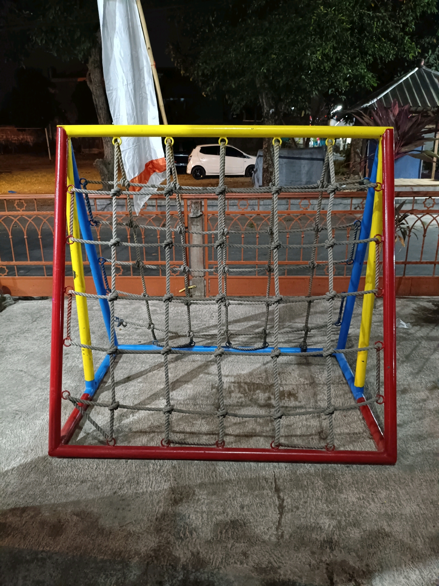 An outdoor shot shows a colorful rope climbing structure in the city of Maguwoharjo, Indonesia. The structure is red, yellow, and blue and is made of metal poles and rope netting. It appears to be a children's playground equipment. In the background, there is a white car, trees, and a fence. The scene is dimly lit, suggesting it might be nighttime. The ground is paved.