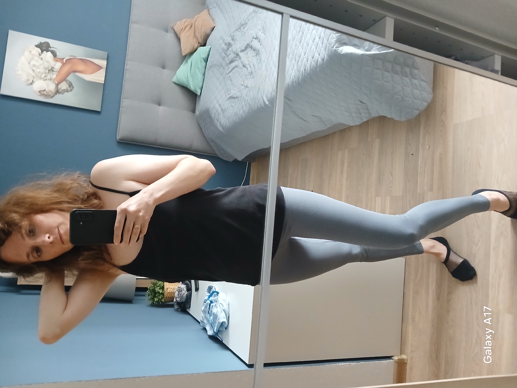 The image is a mirror selfie taken indoors, likely in Yekaterinburg, Russia. A woman with long, curly, reddish-brown hair is captured in the reflection of a large mirror, which appears to be part of a wardrobe. She is wearing a black tank top, light grey ribbed leggings, and black sheer foot socks or low-cut shoes. Her left hand, wearing a ring on the ring finger, holds a black smartphone to take the picture, with her face visible and displaying a neutral or slightly contemplative expression.
The room has light blue walls and a light wooden floor. In the background reflection, a bed with a grey padded headboard and a light blue quilted duvet is visible. Two pillows, one beige and one light mint green, rest on the bed. Above the bed, on the blue wall, hangs a framed piece of art depicting a figure from the back, partially obscured by large white floral elements. To the woman's right in the reflection, on a low white cabinet, there is a small potted green plant in a woven basket and some folded fabric, possibly a light blue and white checkered item. The upper part of the reflection reveals shelves or the interior of a wardrobe. The text "Galaxy A17" is visible on the bottom right corner of the mirror, indicating the phone model used to capture the image. The scene conveys a casual, personal moment in an indoor setting.