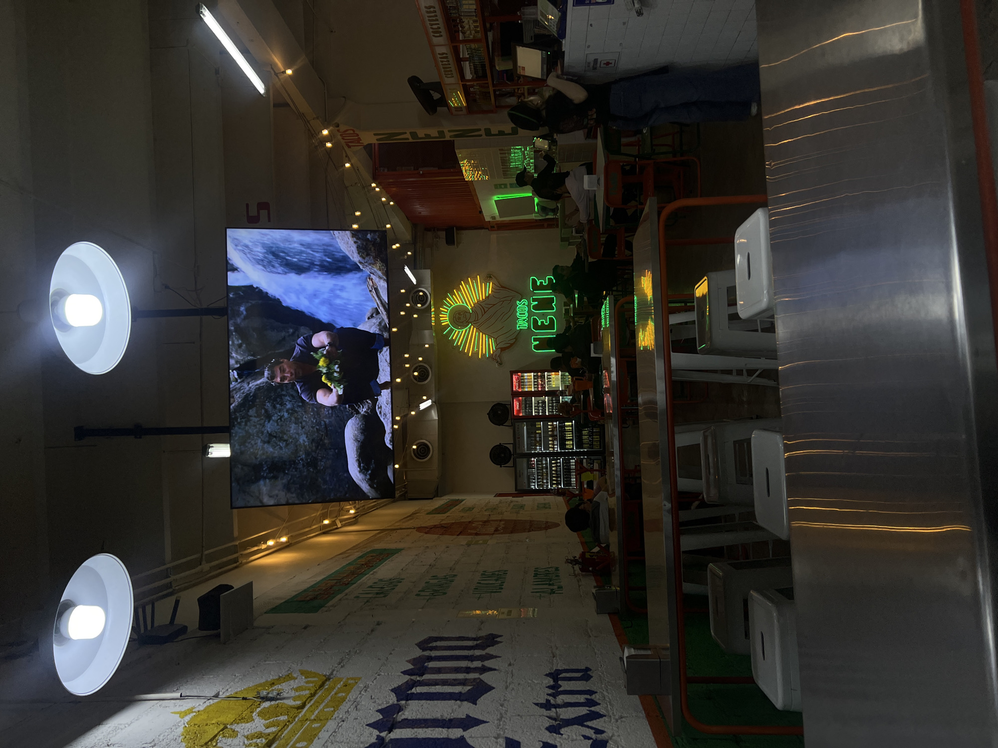 This image captures the interior of a vibrant and casual restaurant or bar, likely in Mexico City. The scene is illuminated by a mix of artificial lights, including bright white task lamps, overhead fluorescent strips, and numerous decorative string lights, suggesting an evening setting.
On the left side, a large wall features striking white murals, prominently displaying "Corona Extra" branding and other Spanish words like "TACOS," "VOLCANES," and "JALAPEÑOS." Two large, circular white pendant lights with exposed bulbs extend from this wall. Above them, a large flat-screen display shows a man holding yellow flowers near a waterfall, adding a dynamic visual element to the space.
Towards the center and right, the area is dominated by a long, metallic counter or bar, with several white bar stools. Behind the bar, a large and colorful neon sign is a focal point, featuring a stylized figure with a halo in yellow and orange neon, alongside the prominent green neon text "TACOS NENE." Further back, shelves are stocked with bottles under signs reading "CERVEZAS" (beers) and "COCTELES" (cocktails), with refrigerators full of drinks below.
Several people are visible throughout the space. Some are seated at the bar or at tables in the background, appearing to be patrons enjoying food and drinks. One person is standing behind the bar, possibly staff. The overall atmosphere is lively and inviting, characterized by the eclectic lighting, modern decor, and prominent food and beverage signage.