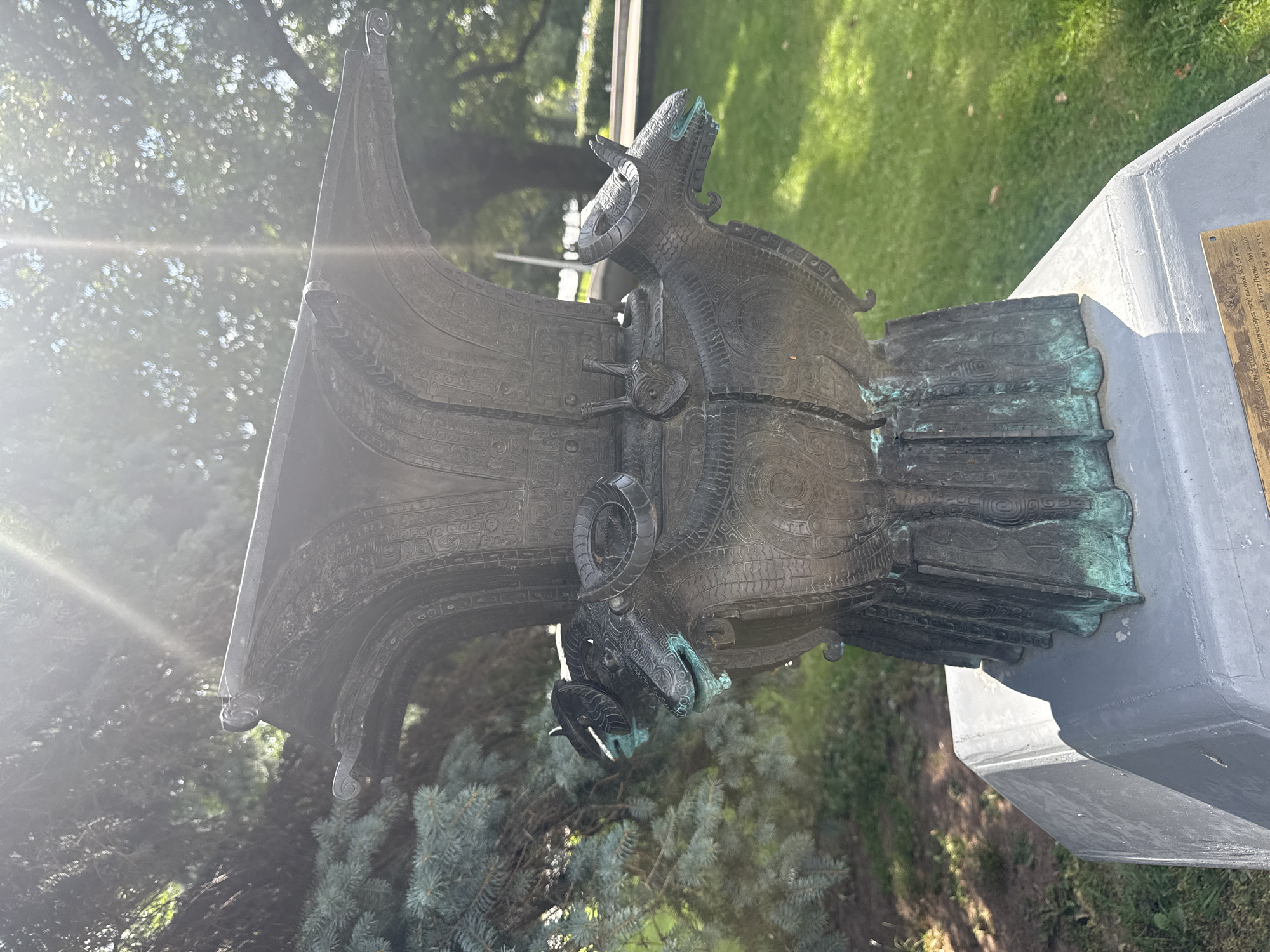 The image shows a bronze sculpture of a vessel with four ram heads at its base, resting on a concrete pedestal. The sculpture is positioned outdoors in a park-like setting, with green grass and trees in the background. Sunlight streams through the trees, creating lens flare and dappled light on the sculpture and surrounding area. The sculpture exhibits intricate detailing, with stylized animal motifs and decorative patterns. The bronze has a weathered appearance, with areas of green patina indicating age. A plaque is visible at the base of the pedestal, though its text is not clearly legible in the provided image. The overall scene suggests an outdoor art installation or monument. The sculpture is located in the city of Mahilyow, Belarus, likely in a public park or square.