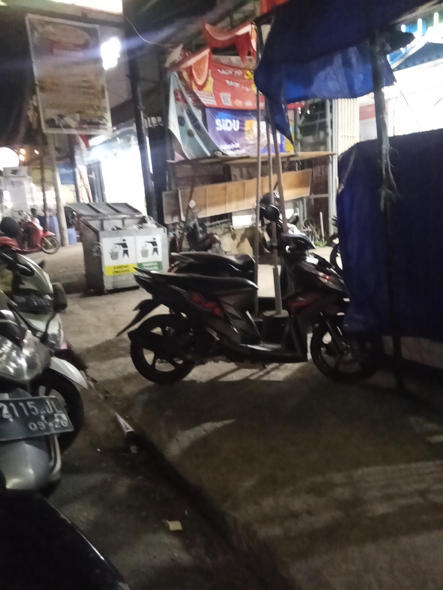 The image depicts an outdoor scene at night, likely in an urban or semi-urban setting. In the foreground, several parked motorcycles are visible. One motorcycle in the lower left is partially shown, revealing a license plate that reads "2115 JL 09-26". Another motorcycle is parked to its right. In the middle ground, there are two large trash bins. The bin on the left has two white icons of a person throwing something into a bin above the words "SAMPAL ORGANIK". The bin on the right has a similar icon and text. Behind the trash bins, a dark-colored motorcycle is parked. Further in the background, there are some structures that appear to be part of a market or a roadside stall, with awnings and hanging banners. One banner in the background has the word "SIDU" on it. The lighting suggests it is nighttime, with artificial lights illuminating the scene. The ground is paved and appears somewhat wet or reflective. The general atmosphere is one of a busy, functional area, possibly a commercial or public space.
