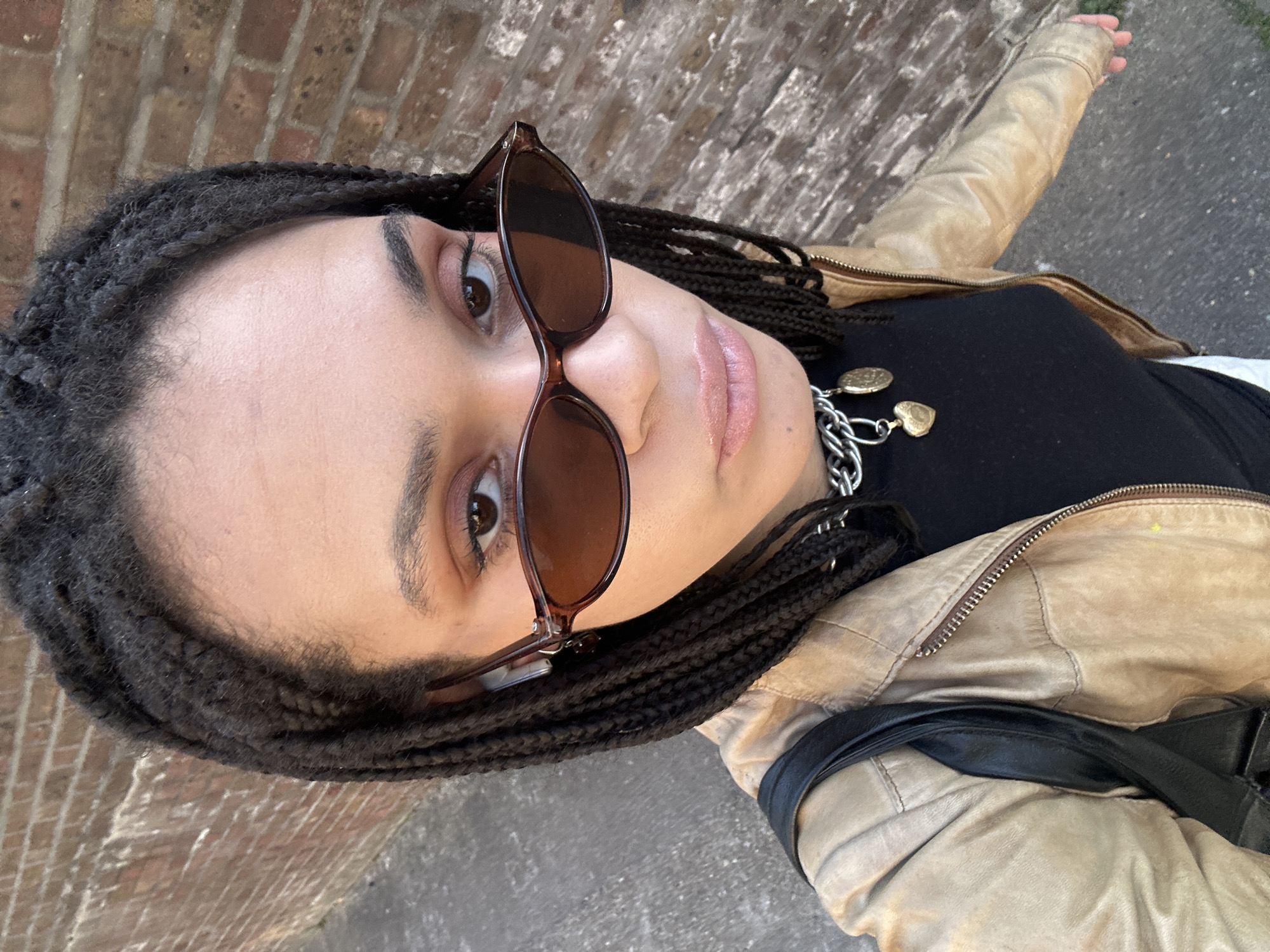 The image presents a close-up, high-angle portrait of a person, likely female, captured in a slightly tilted orientation. The subject has medium-brown skin and dark, thick braided hair, possibly dreadlocks, which frames their face.
They are wearing dark brown, rectangular sunglasses perched on their nose, partially obscuring one eye, while the other brown eye is clearly visible and directed towards the camera. Their eyebrows are dark and well-defined, and their lips are full with a glossy, light pink or nude finish.
The person is dressed in a black top worn under an open, light tan or beige faux-leather jacket. The jacket features a zippered front and what appears to be quilted or ribbed detailing on the shoulders. Around their neck, a chunky silver-colored chain necklace is visible, adorned with three charms: two small, gold-colored heart pendants and one gold-colored, coin-like disc. A black strap, possibly from a shoulder bag, is visible over their right shoulder.
In the background, a reddish-brown brick wall suggests an urban setting, and a paved surface is visible below, likely a sidewalk or street. The lighting appears bright and natural, indicating it is daytime. The person's right arm is extended upwards and outwards, suggesting they might be holding the camera for a selfie. The overall mood is casual and confident. No visible text is present in the image. The location context is London, United Kingdom.