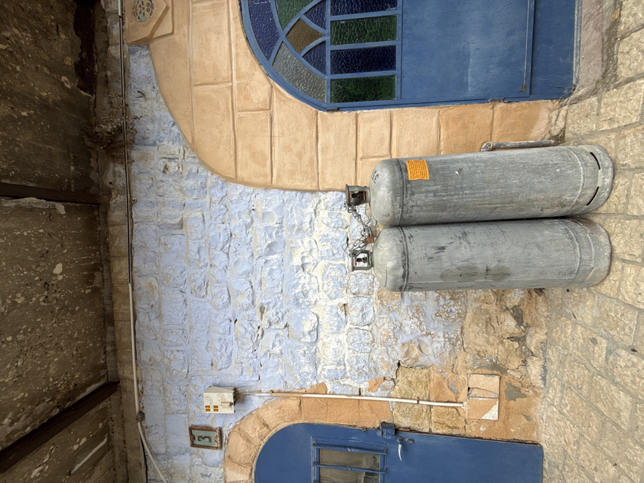 The image depicts an outdoor architectural scene in the city of Safed, Israel, characterized by a weathered stone wall and two doorways. The wall features a rough-hewn stone construction, with the central and upper-left sections painted a light, faded blue. Exposed natural stone in shades of tan and brown is visible in the lower sections and framing the doorways.
Two arched doorways are present. The left doorway is a solid blue door with a small, rectangular window and a horizontal handle, set within a tan-colored arched frame. Above and slightly left of this door, a small framed number "3" is affixed to the blue wall, adjacent to a small, off-white utility box marked with "33" and "30." A white conduit runs vertically downwards from this box. The right doorway is also blue, featuring an arched top section with multiple panes of stained glass in hues of blue, green, and amber. This doorway is also set within a tan-colored arched frame, which includes an ornate, carved or molded decorative element extending upwards and to the right.
In the foreground, two tall, cylindrical, gray metal gas canisters stand upright, leaning against the central part of the wall. They are connected at their tops by a short metal chain. The canister on the right displays a yellow warning label. Both canisters show signs of wear and surface discoloration.
The ground is paved with irregularly shaped, light brown/tan cobblestones or flagstones. Above the wall, a rough, mottled concrete or plaster ceiling extends forward, supported by two dark, possibly wooden or metal beams. The upper wall sections and ceiling show considerable weathering and staining. No people or animals are present in the scene.