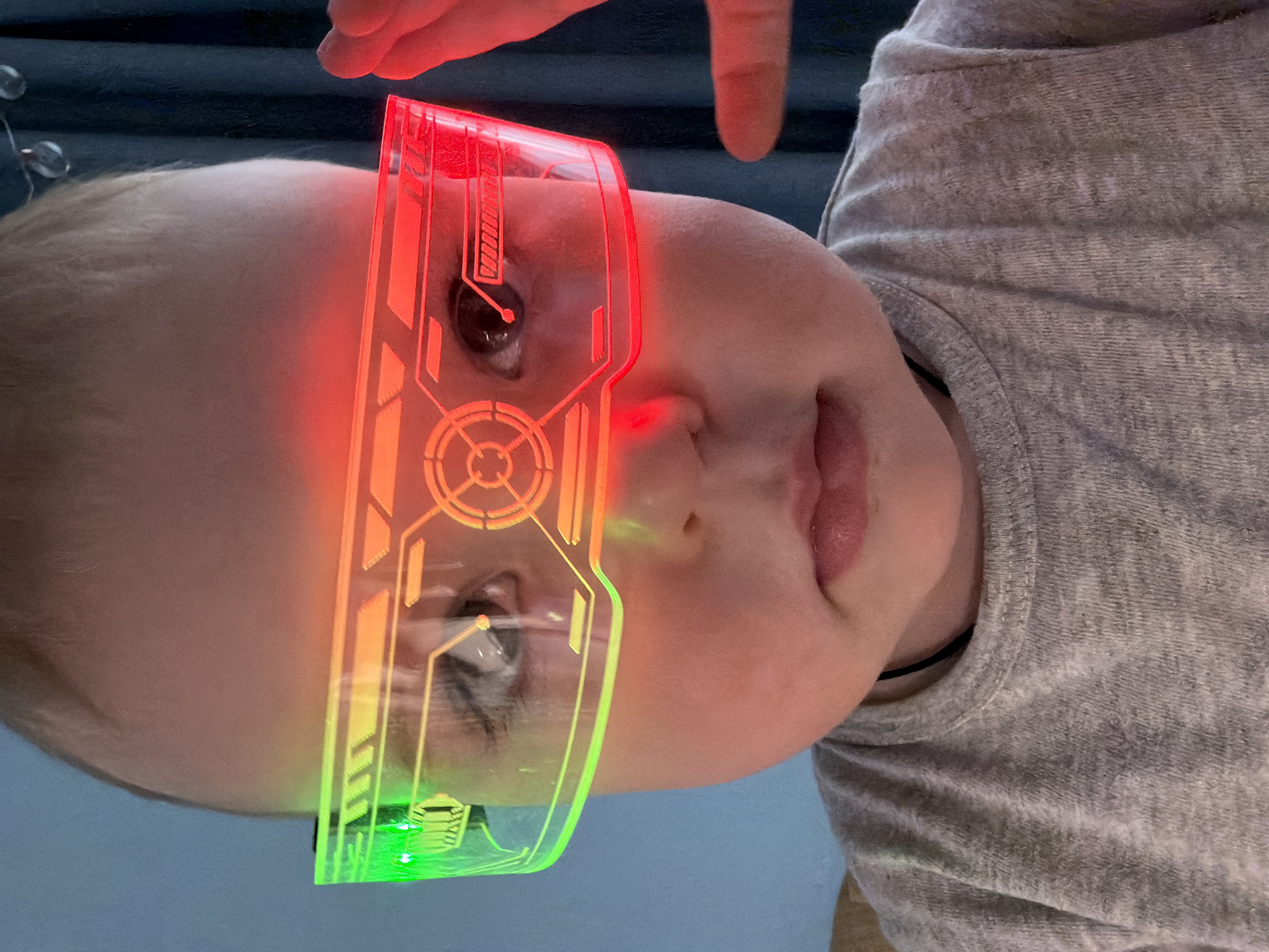 A close-up, portrait-oriented image features a baby or young child, seen from the chest up, with their head slightly turned towards the camera. The child is wearing an illuminated, futuristic-style visor or glasses that cover their eyes. The transparent visor displays an intricate, circuit-board-like pattern, glowing vibrantly with red light on the left side, transitioning through orange-yellow in the center, to green light on the right side. The baby's eyes are visible through the translucent material, looking directly forward with a neutral, slightly curious expression.
The baby has soft, light-colored hair and wears a textured, light gray, possibly fuzzy, garment. An adult's hand, with an index finger extended, is partially visible at the top of the frame, near the baby's forehead, suggesting interaction with the child or the visor. The background is soft-focused; a dark, soft surface, possibly a blanket or cushion, is behind the baby's head, and a lighter, muted blue or gray surface is visible below the baby's face. The scene appears to be an indoor setting, likely with artificial lighting accentuating the glowing visor. The overall impression is one of playful curiosity, with the baby engaging with a whimsical, technological accessory. The location context provided is Votkinsk, Russia. There is no visible text in the image.