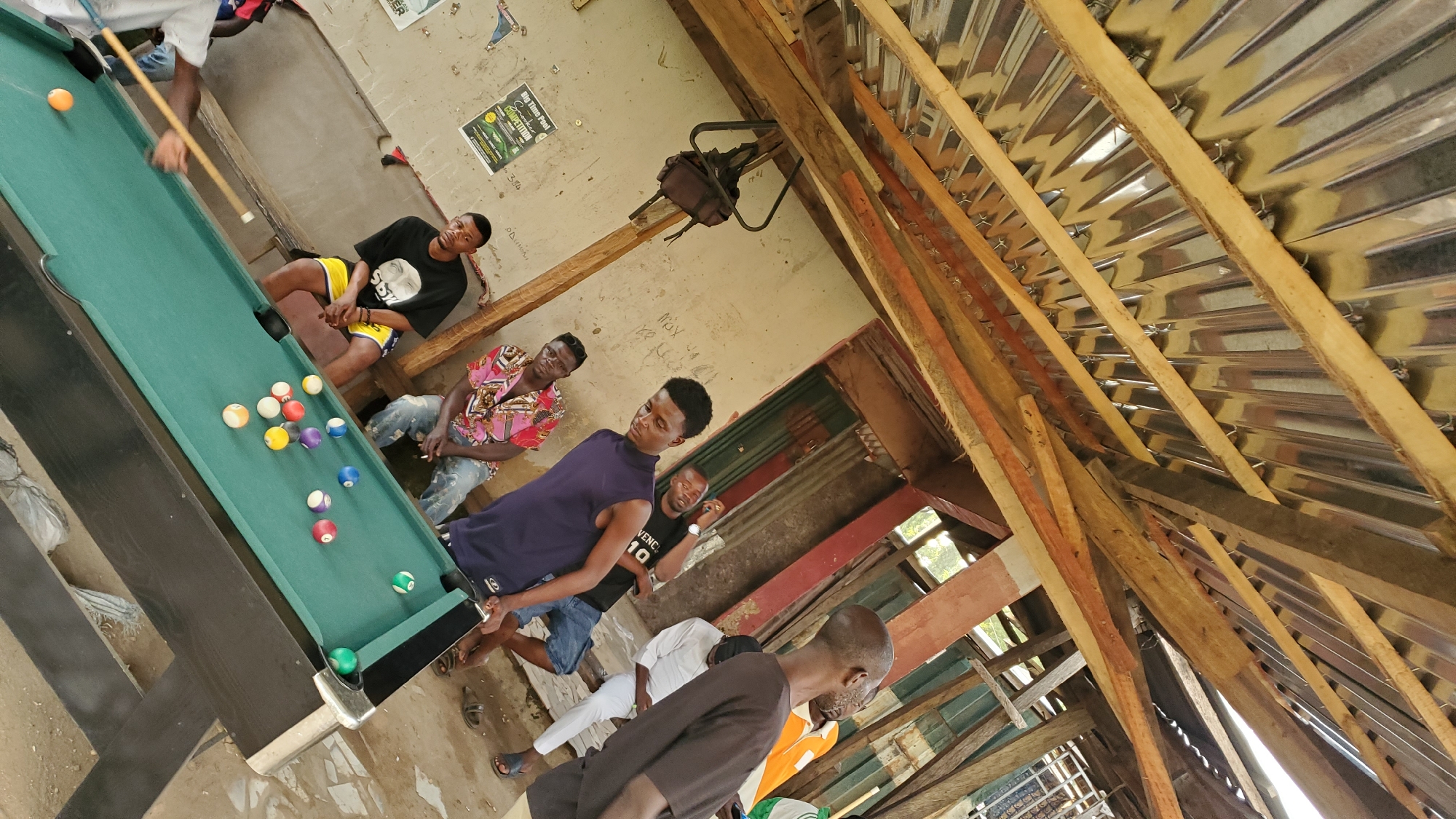 In Jalingo, Nigeria, an outdoor recreational area is depicted, possibly a bar or social gathering spot, characterized by a partially covered corrugated metal roof supported by rough wooden beams. Several young men are present, engaged in or observing a game of pool on a green felt table. One individual in a dark t-shirt and yellow shorts is actively playing, holding a cue stick and aiming at an orange ball. Other men are seated on a bench or standing around the table, some watching the game and others appearing to converse.
The environment is rustic, with concrete flooring and a weathered wall in the background featuring posters. The lighting suggests it is daytime, with light filtering through the open sides of the structure. The overall atmosphere appears casual and social.