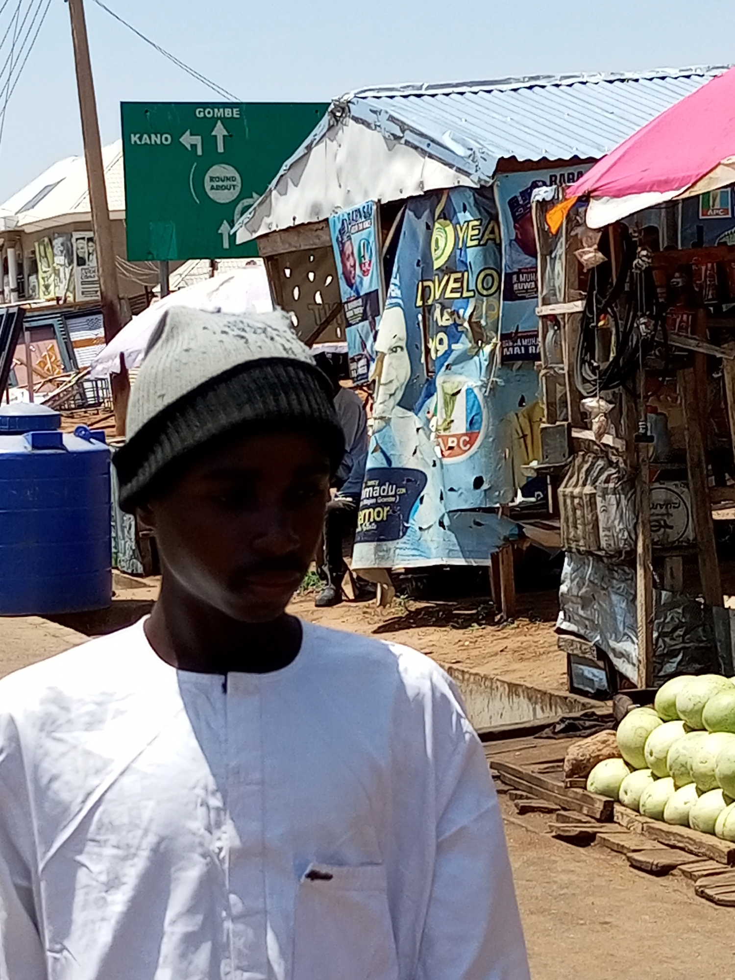 This media file captures a scene in an outdoor market, possibly in Nigeria, given the text on the signage. In the foreground, a young man wearing a white traditional outfit and a knit cap is the primary subject. His face is partially obscured by shadow, but he appears to be looking towards the viewer or slightly off-camera.
Behind the man, the setting reveals a bustling market environment. A prominent green road sign indicates directions to "KANO" with an arrow pointing left and "GOMBE" with an arrow pointing straight up. A circular sign labeled "ROUND ABOUT" is also visible. The market stalls are adorned with colorful posters and banners, many of which appear to be political campaign materials, with images of people and text like "YEA DVELO 19" and "APC." The stalls are covered with corrugated metal and fabric awnings, including a pink one on the right.
Objects in the market include a large blue water tank to the left, wooden crates, and a stacked display of light green fruits, likely watermelons, to the right. The ground is a dusty, reddish-brown dirt. In the background, other people are present, some standing and others walking. The lighting suggests it is daytime, with bright sunlight casting shadows, indicating fair weather. The overall impression is of a lively, everyday market scene.