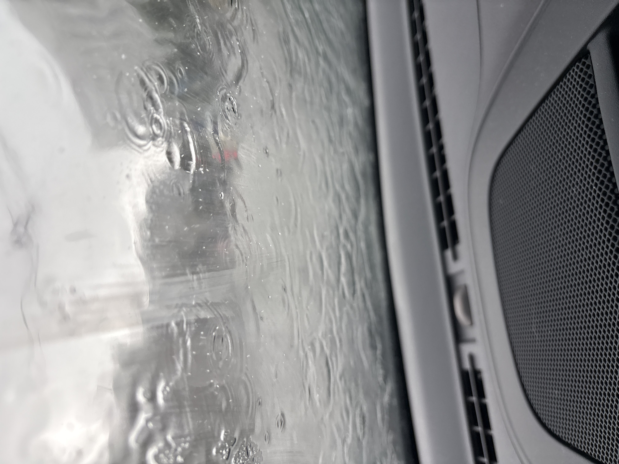 The image shows a close-up of water droplets on a window, likely the interior of a vehicle. The water creates ripples and distorted reflections of the outside environment, suggesting it is raining. Through the water, blurred shapes and colors, possibly parts of other vehicles or buildings, are visible. To the right of the window, a textured speaker grill and part of a car's interior trim are discernible, indicating the vantage point is from inside a car. The overall impression is of being inside a vehicle during inclement weather, observing the rain-streaked view. The context of Vitsebsk, Belarus, suggests a potentially cold or overcast day, consistent with rainy conditions. No text is visible.