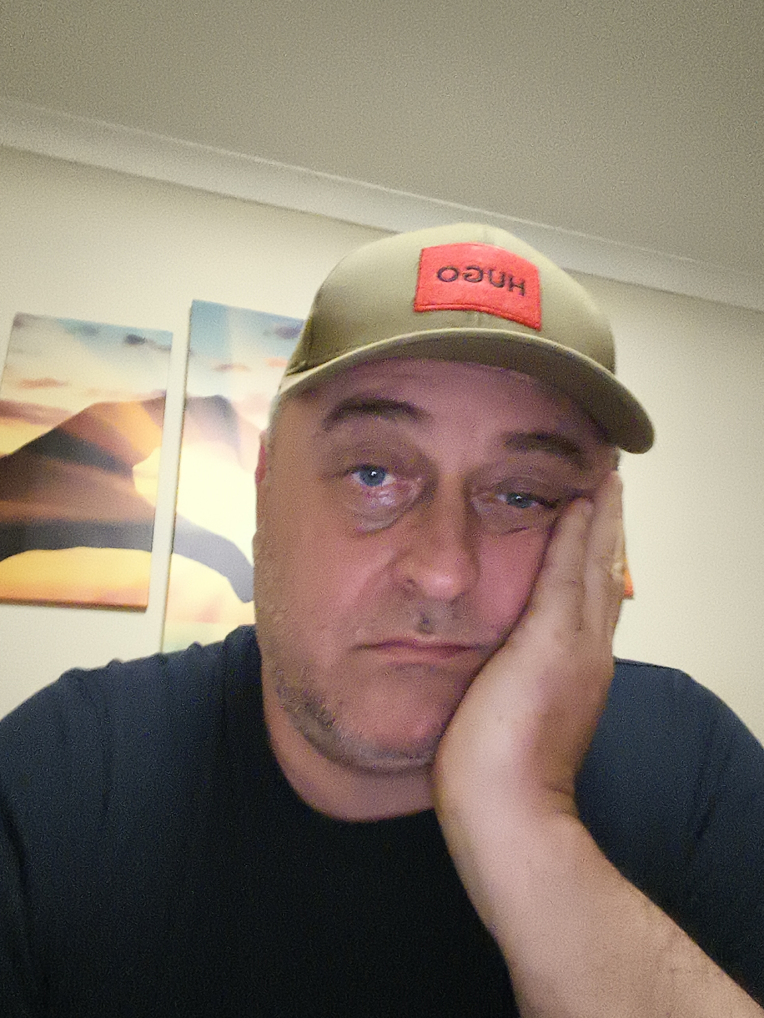 A close-up indoor portrait features a middle-aged man with short, graying hair, wearing a dark t-shirt and an olive green baseball cap. The cap has a red rectangular patch on its front, displaying the inverted white text "OƧUH" (HUGO). The man is seen with his left hand propped against his left cheek and jawline, contributing to his tired, bored, or melancholic expression. His left eye (viewer's right) appears somewhat bloodshot or watery.
The background consists of a plain, light-colored wall, with white ceiling molding visible at the top. To the man's left, two framed artworks or prints are partially visible, depicting abstract landscapes or scenes with warm, muted colors, possibly suggesting sunsets or sunrises. The lighting indicates an indoor setting. This image was captured in Huntingdonshire, United Kingdom.