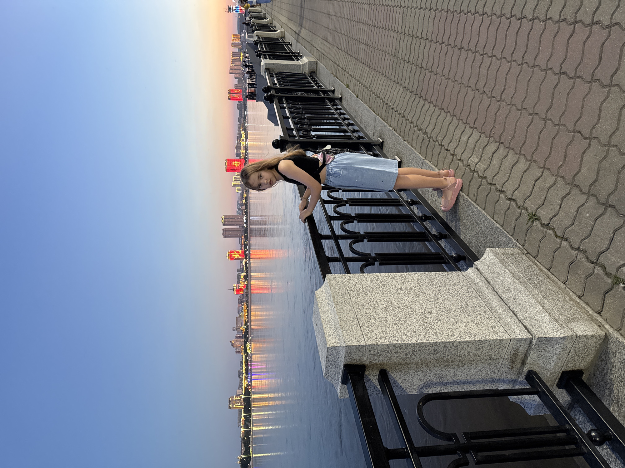 The image depicts a young girl leaning on a black metal railing along a waterfront promenade. She is wearing a black tank top and a light blue denim skirt. Her hair is long and blonde. She is wearing pink slip-on shoes. The promenade is paved with grey interlocking stones. Behind her, across a body of water, is a city skyline illuminated with colorful lights, reflecting on the water's surface. The buildings appear to be residential or office towers. Some of the lights are red and yellow, possibly indicating Chinese flags or decorative lights. The sky is a clear blue with a gradient towards a lighter hue near the horizon, suggesting it is either early morning or late evening, likely sunset given the city lights are on. The overall mood appears peaceful and scenic. The image is oriented vertically, with the girl and railing in the foreground and the city and sky in the background. The location is identified as Blagoveshchensk, Russia.