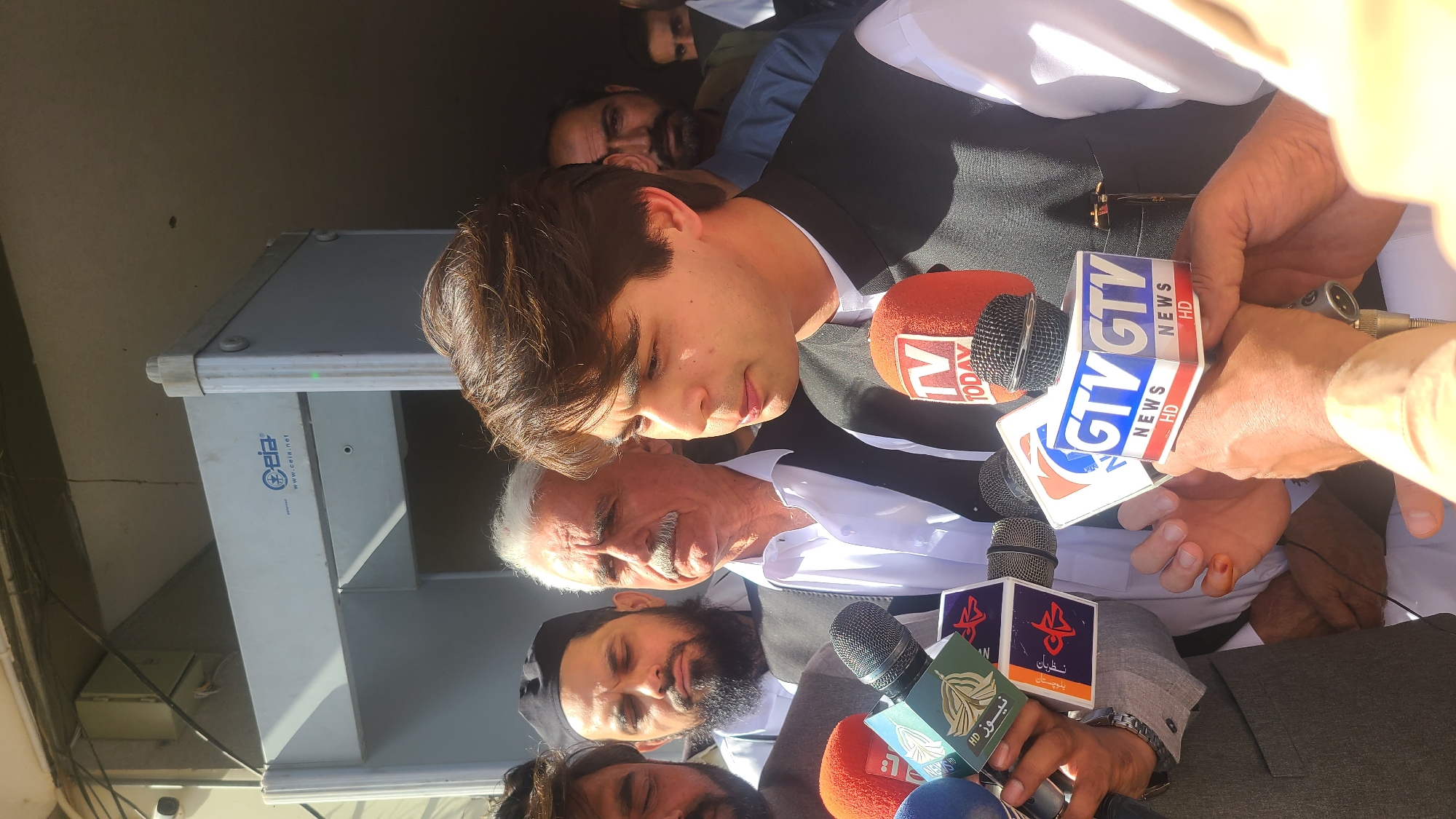 A medium indoor shot shows a young man with short brown hair wearing a white shirt and black vest facing slightly down with several microphones thrust in front of him bearing logos from news networks, including "TV Today" and "GTV News HD," as well as a logo in Arabic script. Behind him is an older man with gray hair and a mustache wearing a white shirt and behind them a man with a beard and a black skullcap. Several other faces are visible in the periphery. The location is possibly Quetta City Tehsil, Pakistan.