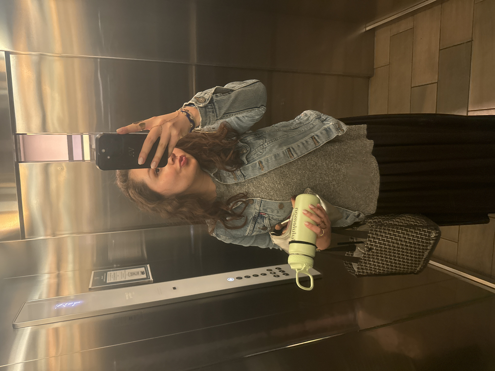 A woman is taking a selfie in an elevator. She is holding a black smartphone in her left hand and a light green water bottle in her right hand. She is wearing a denim jacket, a gray top, and a long black skirt. A patterned bag is on the elevator floor near her feet. The elevator has a stainless steel interior with a control panel visible on the left side. Based on the visible text "somoslulu" on the water bottle, this is likely taken in Mexico City, Mexico, based on the location of the company.
