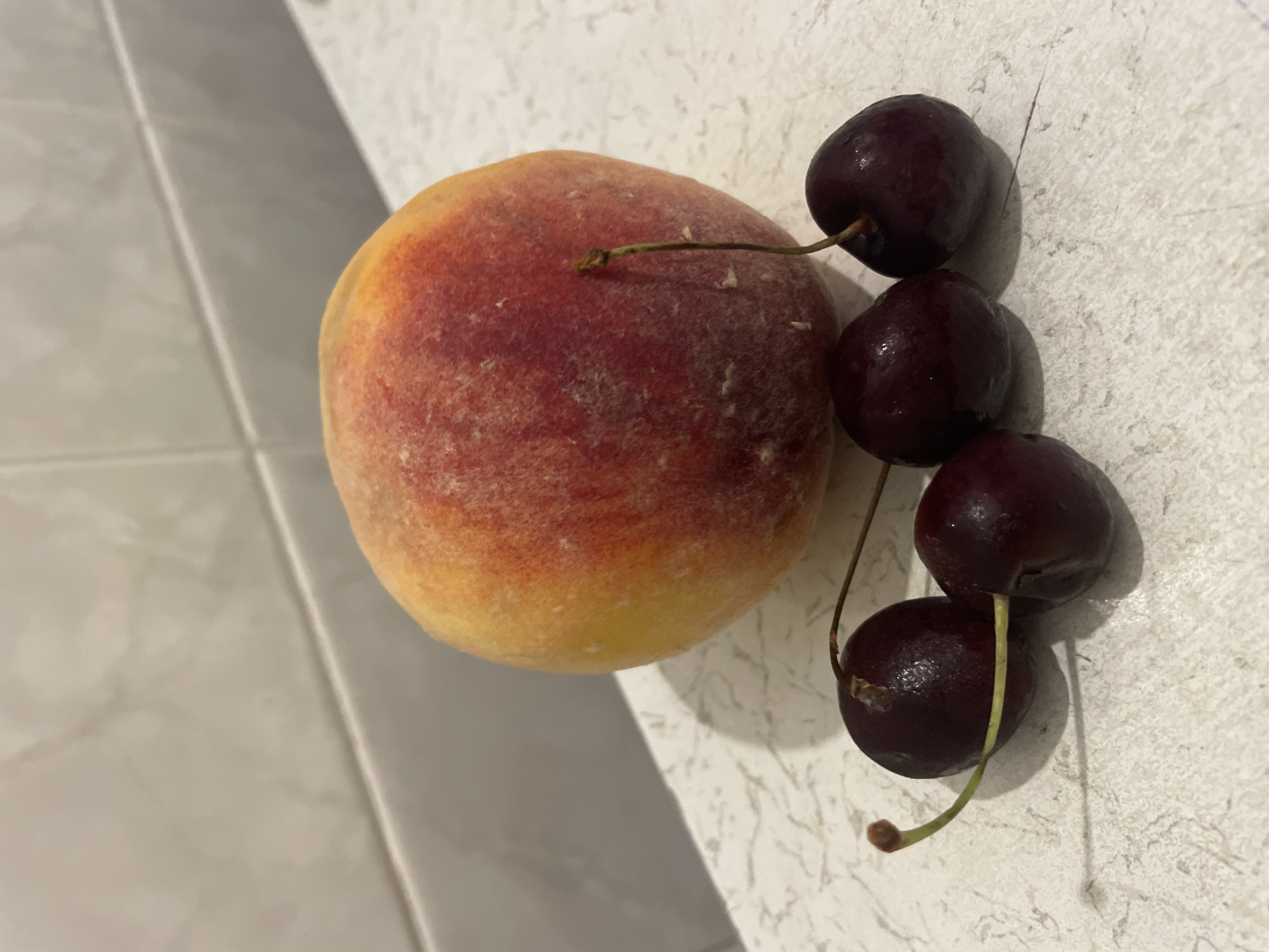 A single peach, a mix of yellow and red, sits on a speckled white countertop. In front of the peach are four dark red cherries, each with a stem. The background is a pale, tiled wall. The scene appears to be indoors, possibly in a kitchen. There is no indication of Kadıköy, Turkey.