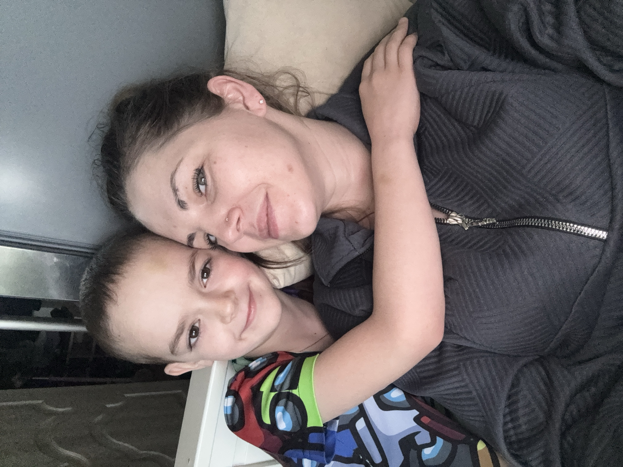 A woman and a child are lying down and embracing in an indoor setting. The woman has fair skin, dark hair pulled back, and facial piercings. She is wearing a dark gray jacket with a zipper. The child, presumably a boy, has short brown hair and is wearing a colorful patterned shirt with blue, green, red, and black elements. They are both smiling at the camera. The background includes a light-colored pillow or cushion and a window or reflective surface. The location is identified as Раздолье, Russia.