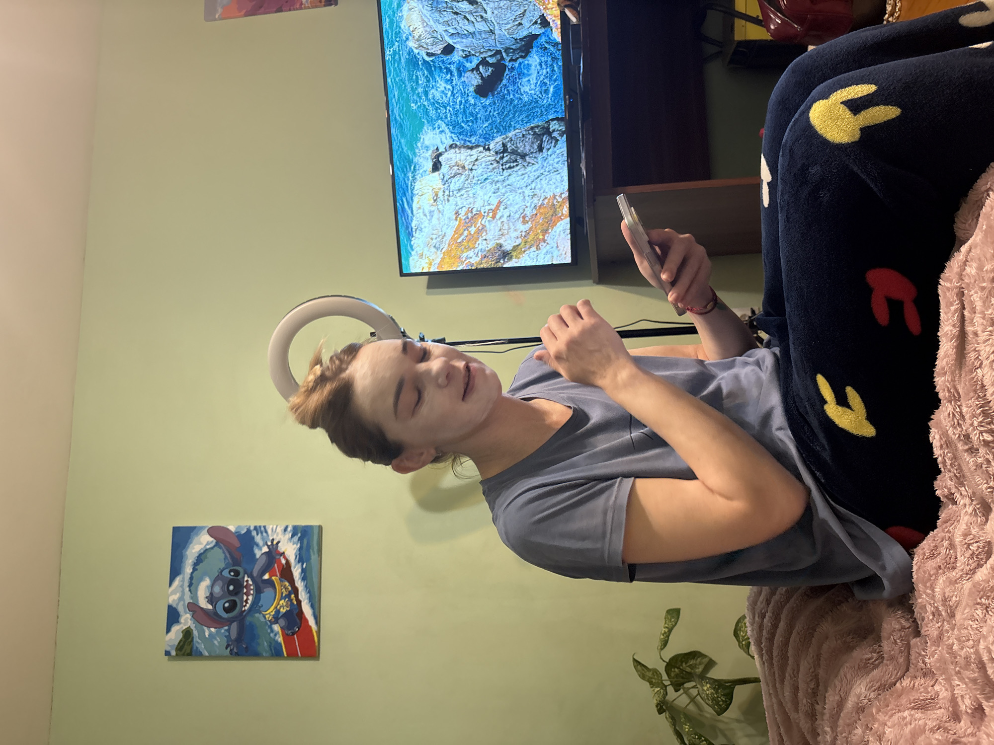 A person with short hair, wearing a grey t-shirt and patterned blue pants, is sitting on a pink blanket, looking at a cell phone. The person's face appears to have some kind of mask or treatment on it. A ring light is positioned above them. Behind the person is a wall painted a light green color, with a television displaying a blue seascape mounted on the wall. Below the television is a dark-colored cabinet. A painting depicting Stitch surfing is on the wall. A potted plant is near the bottom right of the image. The city of Sinyavino, Russia is not discernible from the image.