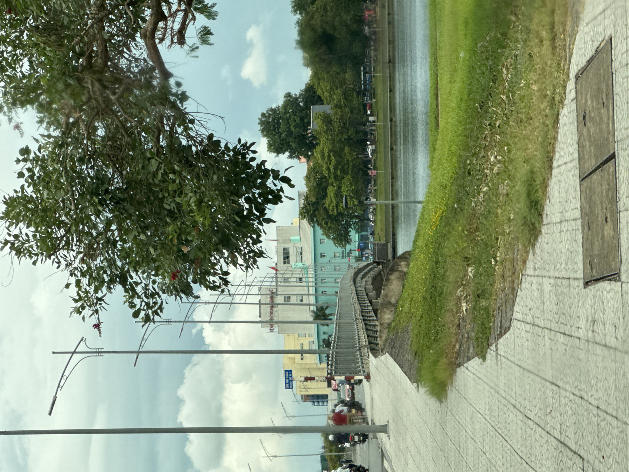 The image depicts a scene in Thuận Hóa Ward, Vietnam. A tiled pedestrian walkway runs along the left side of the frame, bordering a grassy embankment and a body of water on the right. Several lampposts line the walkway. In the background, across the water, is a city skyline with various buildings, including a multi-story building with the text "BỆNH TRUNG ĐỘNG" visible on its facade. A bridge arches over the water in the mid-ground, connecting the two sides of the walkway. Several individuals are present, some on motorcycles and bicycles, navigating the walkway and a nearby road. The sky is overcast with clouds. The environment appears to be a developed urban area adjacent to a waterway.