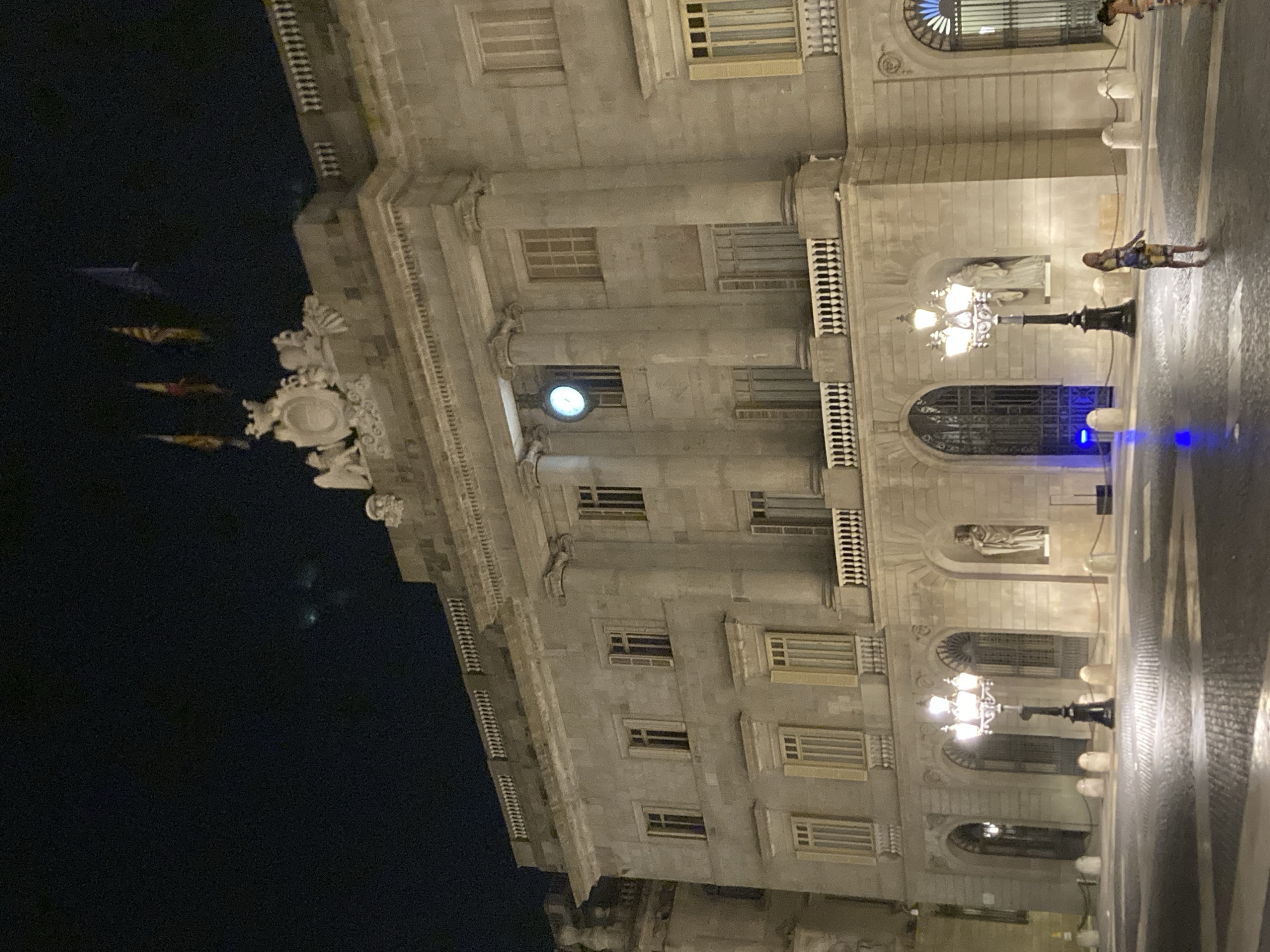 The image shows the Palau de la Generalitat in Plaça Sant Jaume, Barcelona, Spain, at night. The building is illuminated by streetlights and architectural lighting. A clock is visible on the facade. Two women are walking in the foreground. The ground is paved with cobblestones. There are street lamps and bollards. Flags are flying from the roof of the building.