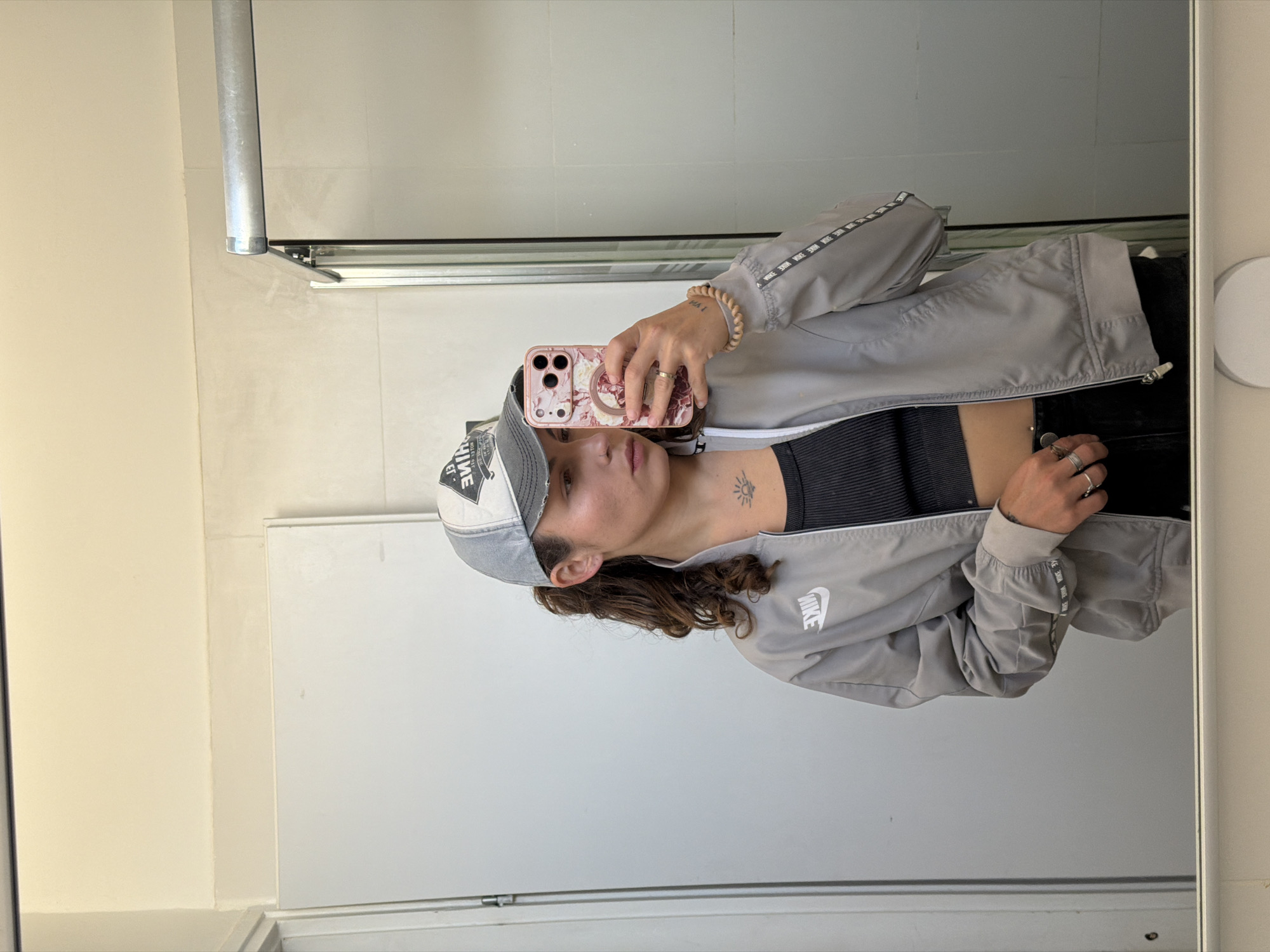 A person is taking a selfie in front of a mirror. The individual is wearing a baseball cap, a grey Nike jacket, and a black top. The person is holding a phone with a floral patterned case. The environment appears to be a bathroom with white walls. There is no verifiable information to confirm a specific location in Kiryat Bialik, Israel.
