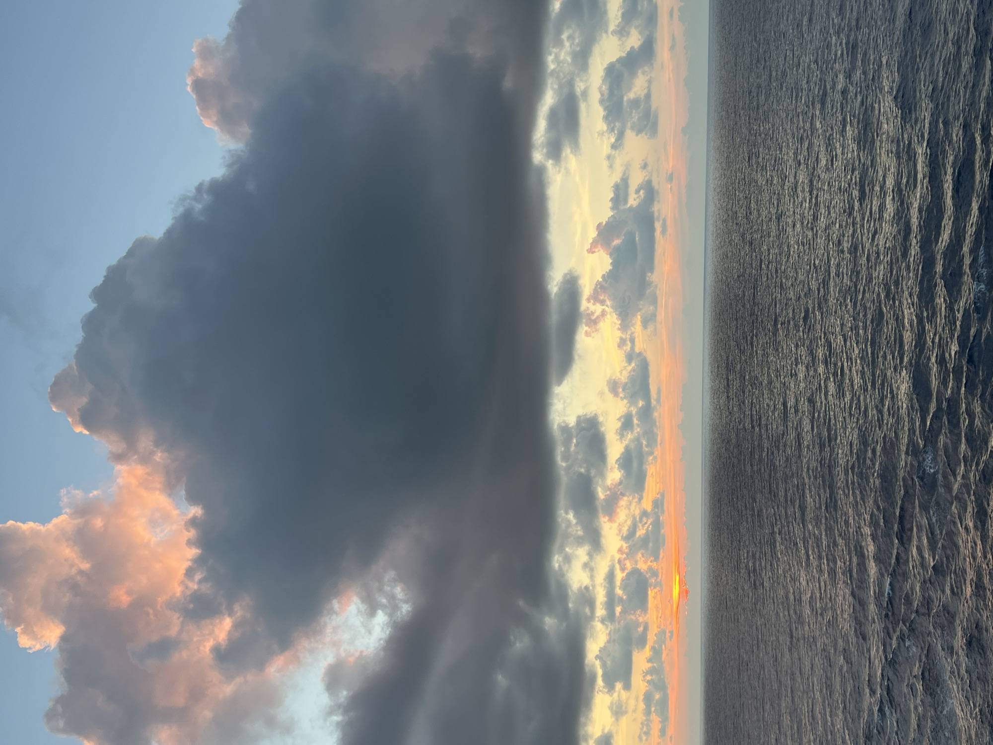 An image displays a vast expanse of dark, choppy ocean water meeting a horizon under a sky painted with dramatic clouds. The clouds are a mix of deep grays and lighter shades tinged with pink and orange, suggesting either sunrise or sunset. A bright, fiery orb, partially obscured by clouds, sits low on the horizon, casting a warm glow on the clouds and the surface of the water. The scene is devoid of any landmass, structures, or individuals, indicating an open-water environment. The perspective suggests the viewer is on a ship or elevated platform overlooking the sea.