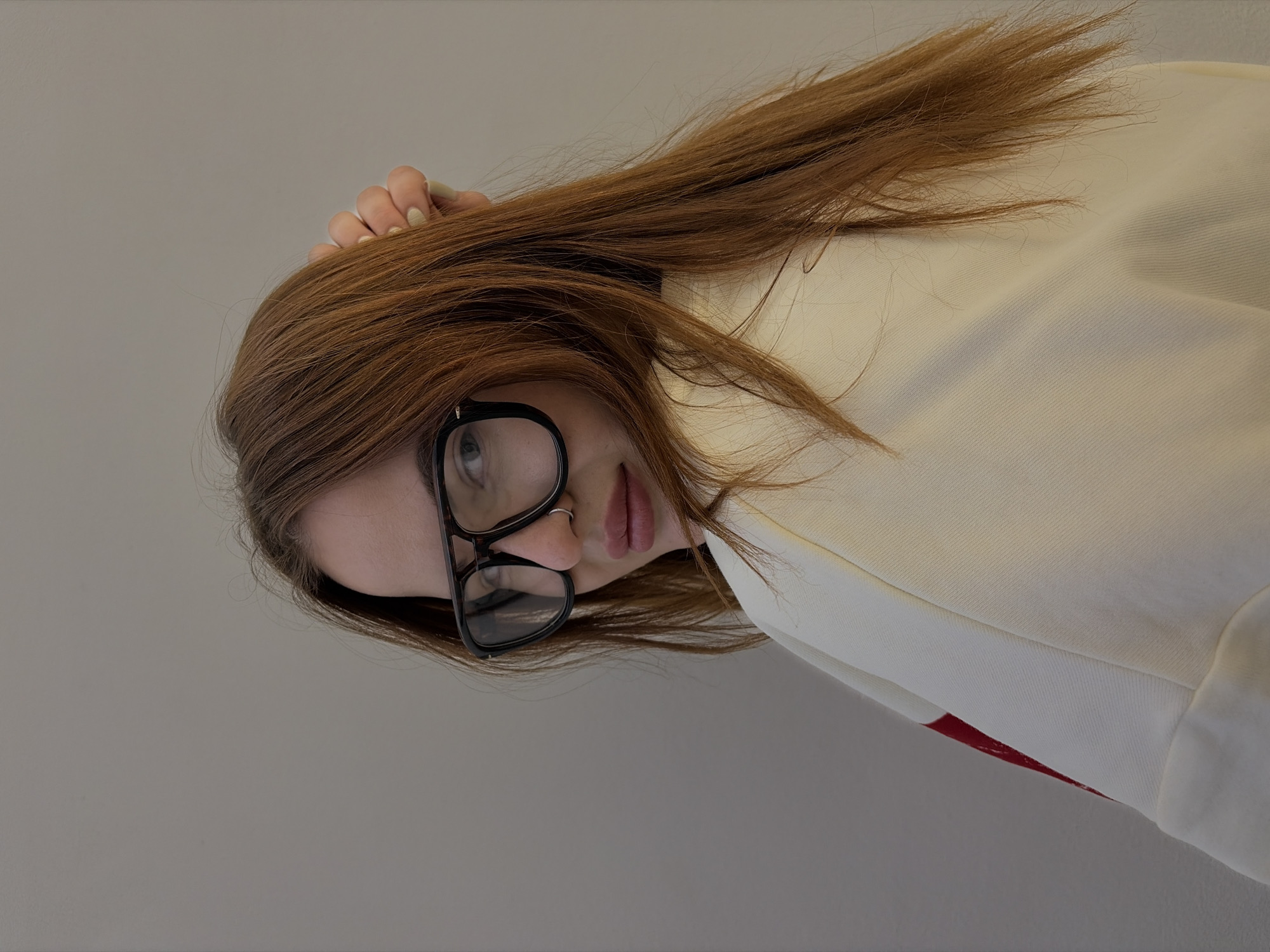 The image presents a sideways portrait of a young female individual against a plain, light grey background. Her head is positioned on the left side of the frame, with her body extending to the right. She has long, straight, reddish-brown hair that spreads towards the top and right of the image. Her right hand, adorned with light green/gold manicured nails, is partially visible, seemingly resting in or holding her hair near the top of her head.
The subject wears large, black-framed glasses that obscure much of her eyes, and a silver septum piercing is visible on her nose. Her lips are slightly parted, suggesting a neutral or softly thoughtful expression. She is dressed in a light-colored, possibly cream or off-white, long-sleeved top or jacket. A hint of red fabric is visible beneath the sleeve on the far right.
The individual appears to be posing for the camera, with her gaze directed upwards through her glasses. The setting is indoors, indicated by the uniform lighting and plain wall, with no specific location cues other than the provided context of Krasnodar, Russia. No text is visible in the media file.