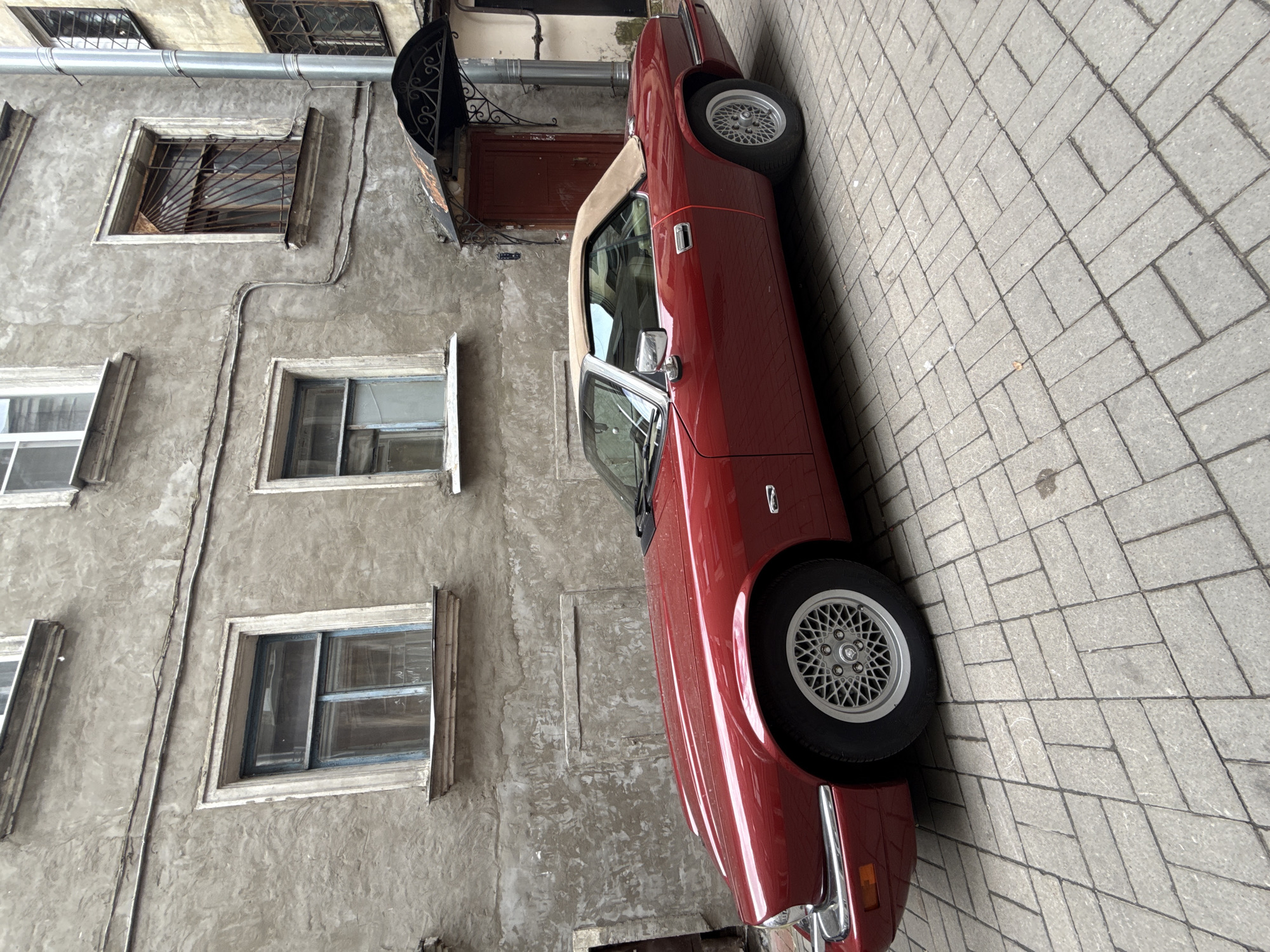 A red Jaguar XJS convertible is parked on a cobblestone alleyway in front of a weathered, grey, multi-story building. The building has multiple windows with old wooden frames and some have metal grates. An arched, decorative metal canopy with peeling paint is mounted above a dark red door. The sky is overcast. The car's top is down, revealing a tan interior. The wheels are silver alloy with a distinctive spoke pattern. The scene is located in Saint Petersburg, Russia.