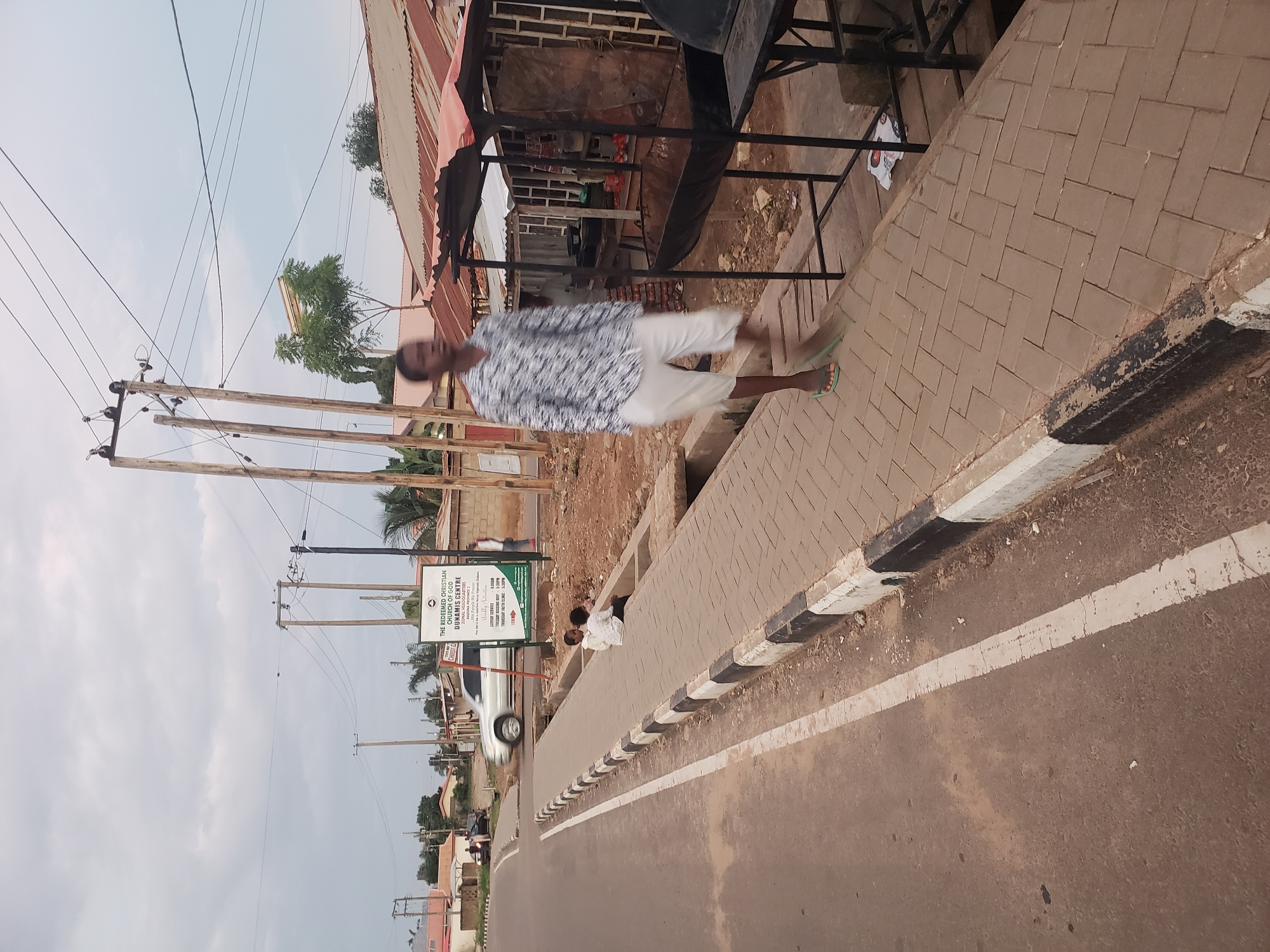 The image depicts a street scene in Narayi, Nigeria, with a focus on a paved walkway adjacent to a road. In the foreground, a young man in a patterned shirt and white shorts is walking on the paved walkway, captured in motion. To the left, a road with white lane markings is visible. Two children are seated on the edge of the walkway. In the background, there are buildings with corrugated metal roofs, utility poles with power lines, and a sign for "THE REDEEMED CHRISTIAN CHURCH OF GOD DUNAMIS CENTRE". A silver SUV is driving on the road in the middle ground. The environment appears to be an urban or semi-urban setting under an overcast sky.