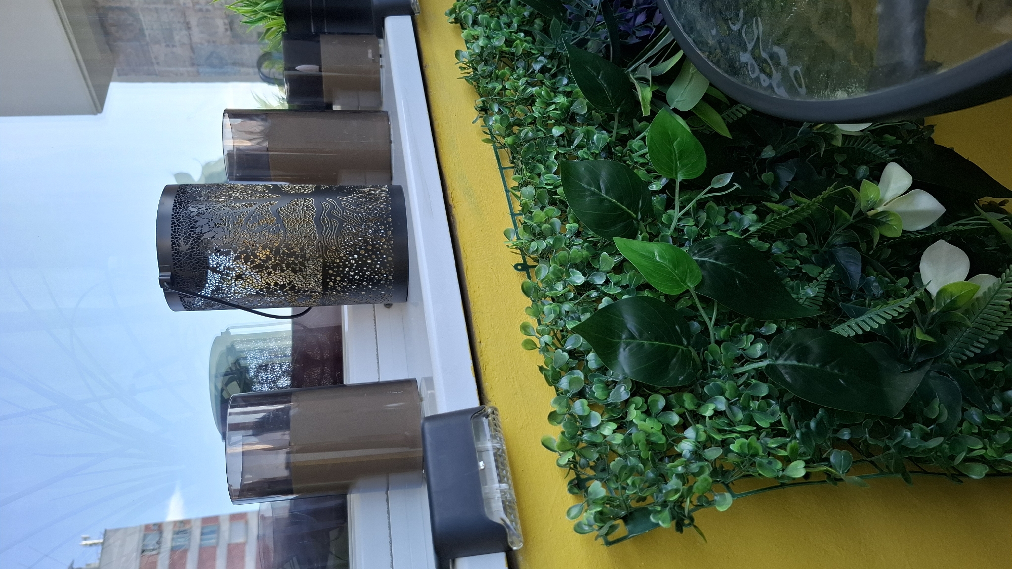 The image shows a close-up of a shopfront display in Sofia, Bulgaria. The building across the street is visible in the reflection of the shop window. There is a white window frame with mounted black metal candle holders and translucent brown sleeves around the candle holders. Next to the window frame is a vertical yellow painted surface, covered in artificial green foliage with a variety of leaf sizes and textures, including small round leaves and larger glossy leaves. There are also several white flowers. A small portion of a round black mirror is visible on the top right.