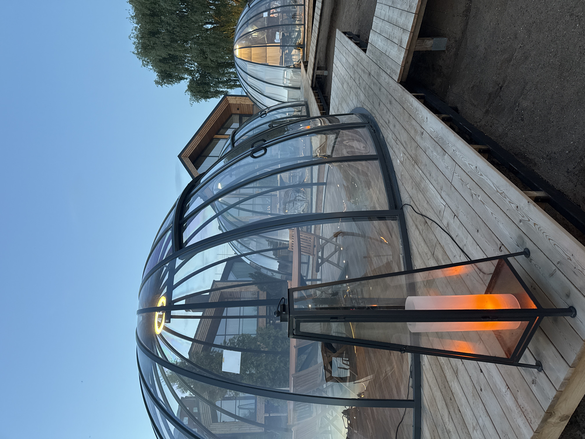 This detailed image captures an outdoor dining or leisure area featuring multiple transparent geodesic dome structures at dusk in Votkinsk, Russia. The scene is illuminated by internal and external light sources under a clear, transitioning twilight sky.
In the foreground, a tall, black metal lantern with glass panels stands on a wooden platform, housing an upright, cylindrical, amber-glowing artificial candle. This platform extends into a wooden walkway connecting several of the dome structures.
Three prominent dome structures are visible. The two closest domes, positioned centrally and to the left, consist of dark, segmented frames enclosing clear, transparent panels. Through these, multiple empty wooden tables and chairs are visible, along with subtle interior string lighting. The dome furthest to the right is also transparent but shows brighter interior illumination and white curtain-like elements, enclosing additional tables and chairs. All domes are elevated on light-colored wooden decking.
Behind the domes, a contemporary building with distinctive wooden cladding and large glass windows is visible, featuring a prominent dark-framed, triangular roofline. To the right of the scene, a large, leafy tree stands against the deepening blue sky. The ground beneath and around the wooden platforms is dark, unpaved earth or gravel. No individuals are present in the image.