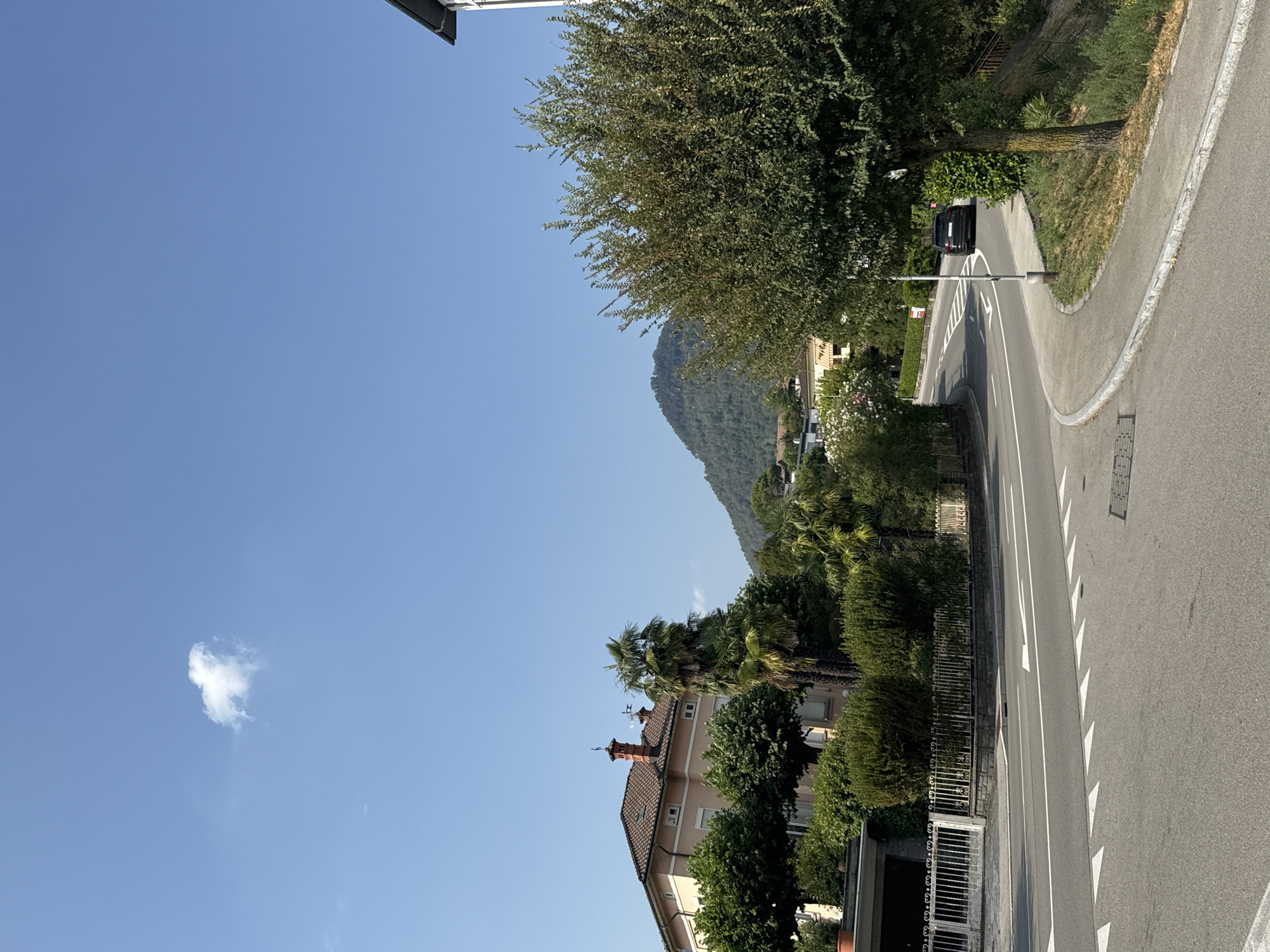 The image shows a street scene in Morbio Inferiore, Switzerland. A paved road curves through a residential area with houses and lush vegetation. A dark-colored car is seen driving away on the road. The sky is clear blue with a single small white cloud. A mountain covered in trees is visible in the background.
