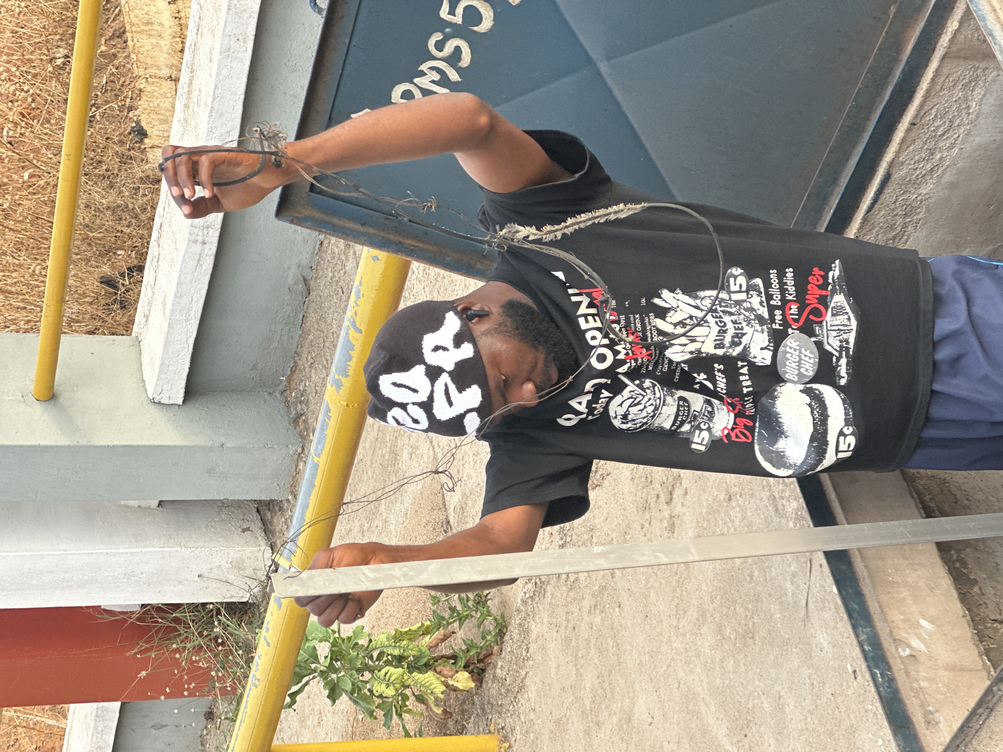 A man with dark skin and a beard is pictured outside near some buildings and infrastructure. He is wearing a black T-shirt with a graphic design, a dark beanie with white lettering, and blue shorts. He is holding a thin, silvery metal bar, and some tangled wire. The image contains partial views of painted yellow railings, concrete structures, and a portion of a blue wall with white lettering that appears to read "PMS 5." There are also patches of vegetation visible near the ground. The setting seems to be an urban or industrial outdoor space. The geographical location of Jos, Nigeria, cannot be verified from this image alone.