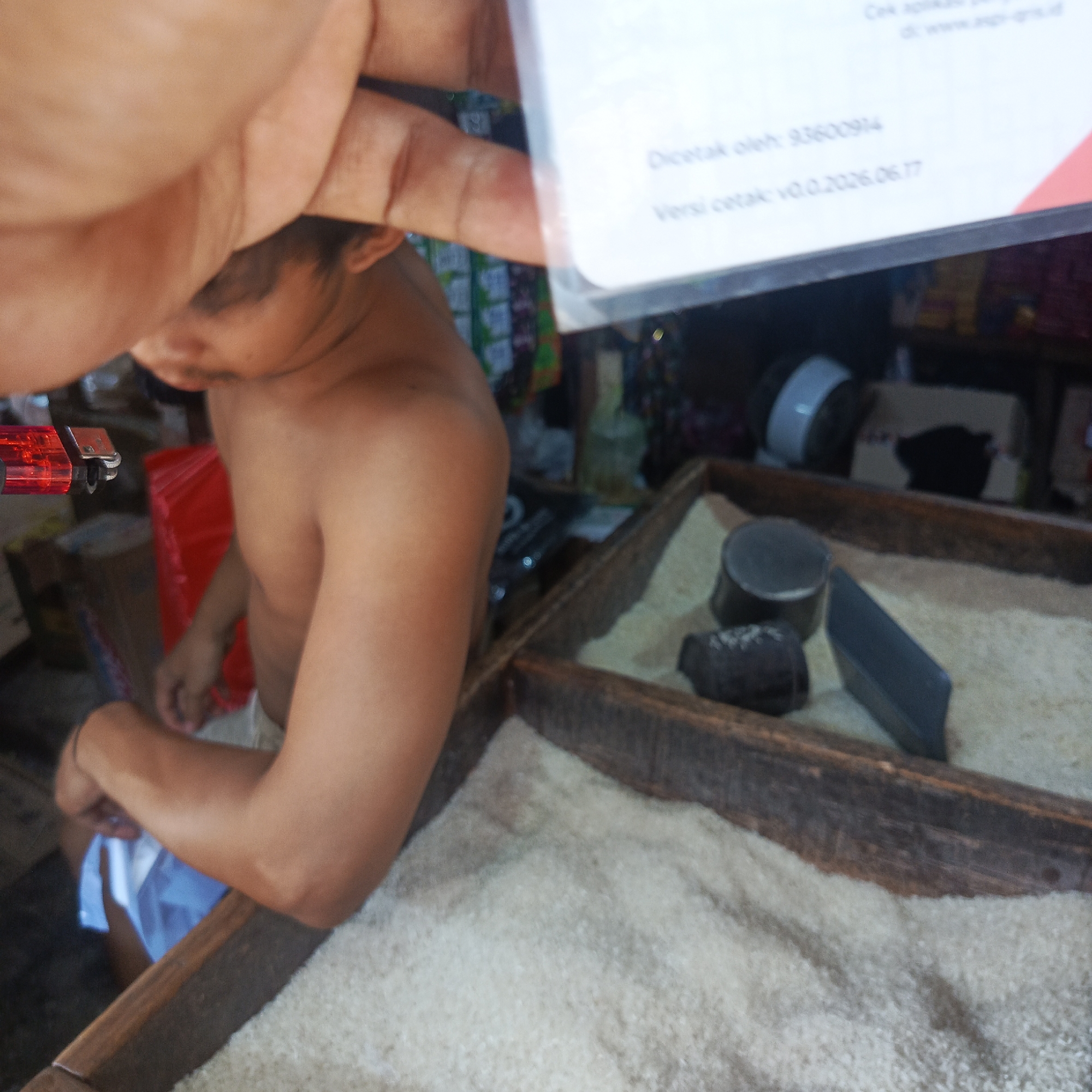 The image shows a close-up view of a person, partially obscured by a hand holding a document. The person appears to be a young male, shirtless, with dark hair, and is looking down. He is wearing a white cloth around his waist. The setting seems to be an indoor market or shop, with shelves of goods visible in the background.
In the foreground, there is a wooden tray filled with rice. Several objects are placed on top of the rice, including a metal container, a dark object that might be a tool, and a flat, dark rectangular object. A red object, possibly a lighter or some kind of tool, is also visible on the left side of the frame.
The document being held in the foreground has some visible text. The text is in Indonesian and includes phrases like "Dicetak oleh: 93600914" (Printed by: 93600914) and "Versi cetak: v0.0.2026.06.17" (Print version: v0.0.2026.06.17).
The lighting suggests it is daytime, but the specific time or weather conditions are not discernible. The overall impression is a candid shot within a local marketplace environment.