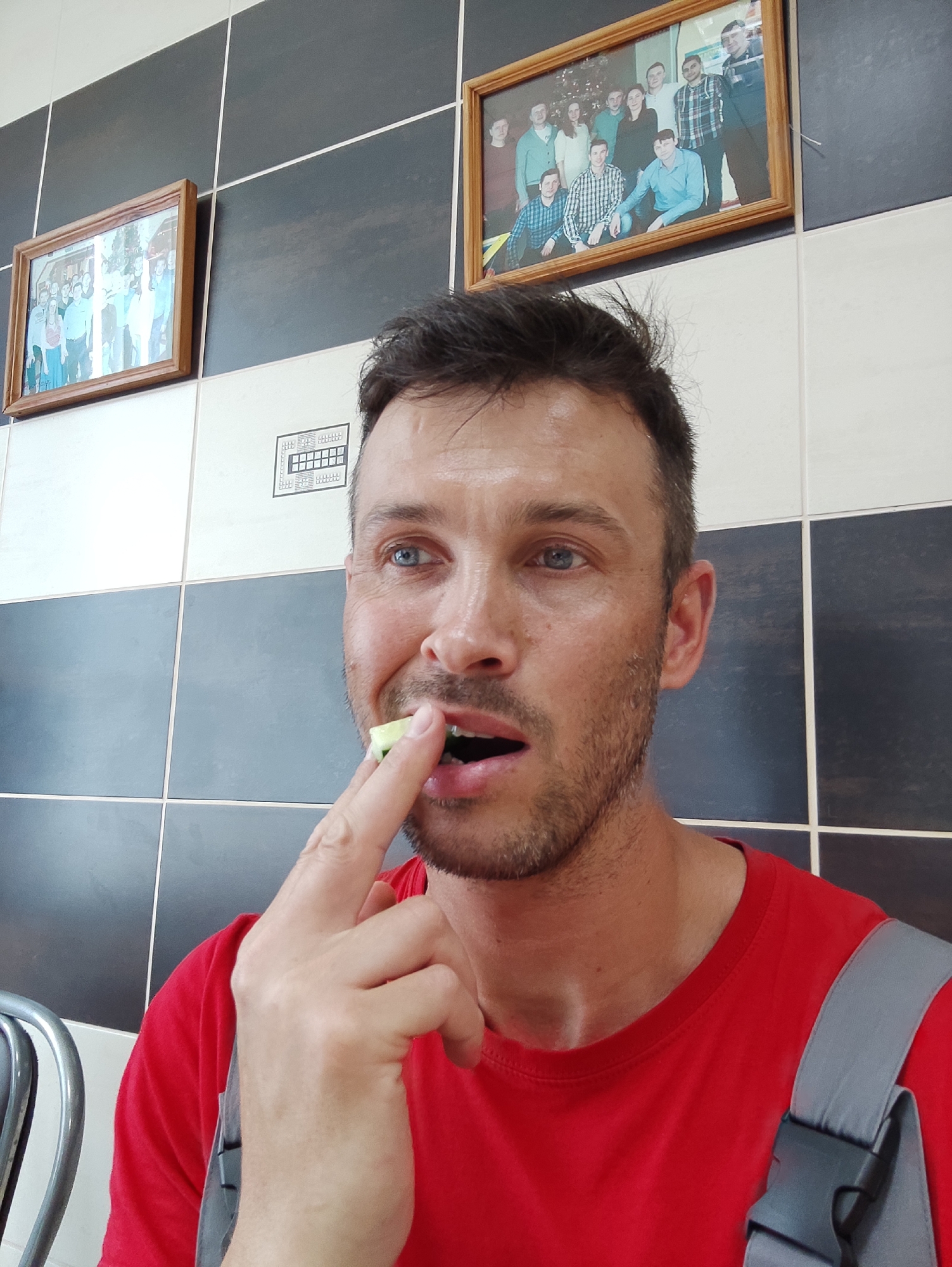 The scene, located in the city of Unknown, Unknown, captures an adult male in an indoor setting. The man, with short dark hair, light blue eyes, and light stubble, occupies the majority of the foreground. He wears a red short-sleeved t-shirt and a grey strap, consistent with a backpack or shoulder bag, is visible over his right shoulder. His left hand is raised, with his index finger and thumb holding a small, light-green, rectangular food item, possibly a piece of cucumber, to his slightly open mouth. His gaze is directed upwards and to the viewer's left. The background consists of a wall tiled in a checkerboard pattern of alternating dark grey/black and white/light grey squares. Two framed wooden pictures are mounted on the upper dark tiles. The picture on the left depicts a group of people. The picture on the right, also showing a group of people, is more clearly visible. Between the man's head and the right picture, a black schematic diagram or chart, resembling a grid of squares, is printed on a white tile. A silver-colored metal chair back is partially visible on the far left of the frame.
