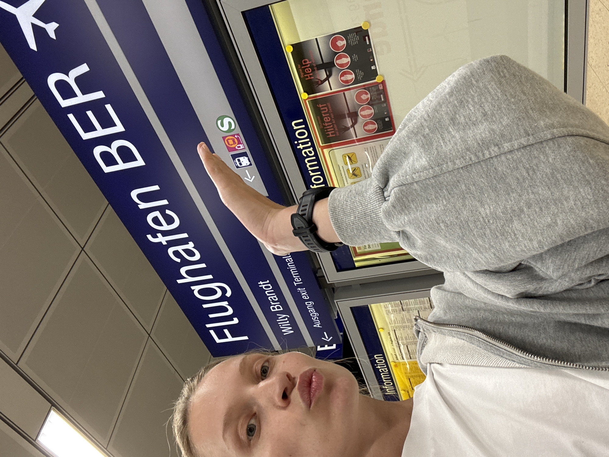 A fair-skinned individual, likely female, with blonde hair pulled back, takes a selfie indoors at Berlin Brandenburg Airport (BER) in Schönefeld, Germany. She is wearing a white t-shirt and a grey zipped sweatshirt, with a black digital watch on her left wrist. Her right hand points upwards towards a prominent blue overhead sign with white text that reads "Flughafen BER" and includes an airplane silhouette. Below, the sign also displays "Willy Brandt" and "Ausgang exit Terminal" with a left-pointing arrow, along with icons for a train, bus, and an S-Bahn (green 'S' circle). Below this main sign, two large framed information displays are visible. The left display is labeled "Information". The right display features two posters attached with yellow push-pin-like elements; one poster is titled "Stummer Hilferuf" (Silent Cry for Help) with the DB logo, and the other is titled "Signal for Help," both illustrating hand gestures for distress. The ceiling is a tiled false ceiling with recessed fluorescent lighting.