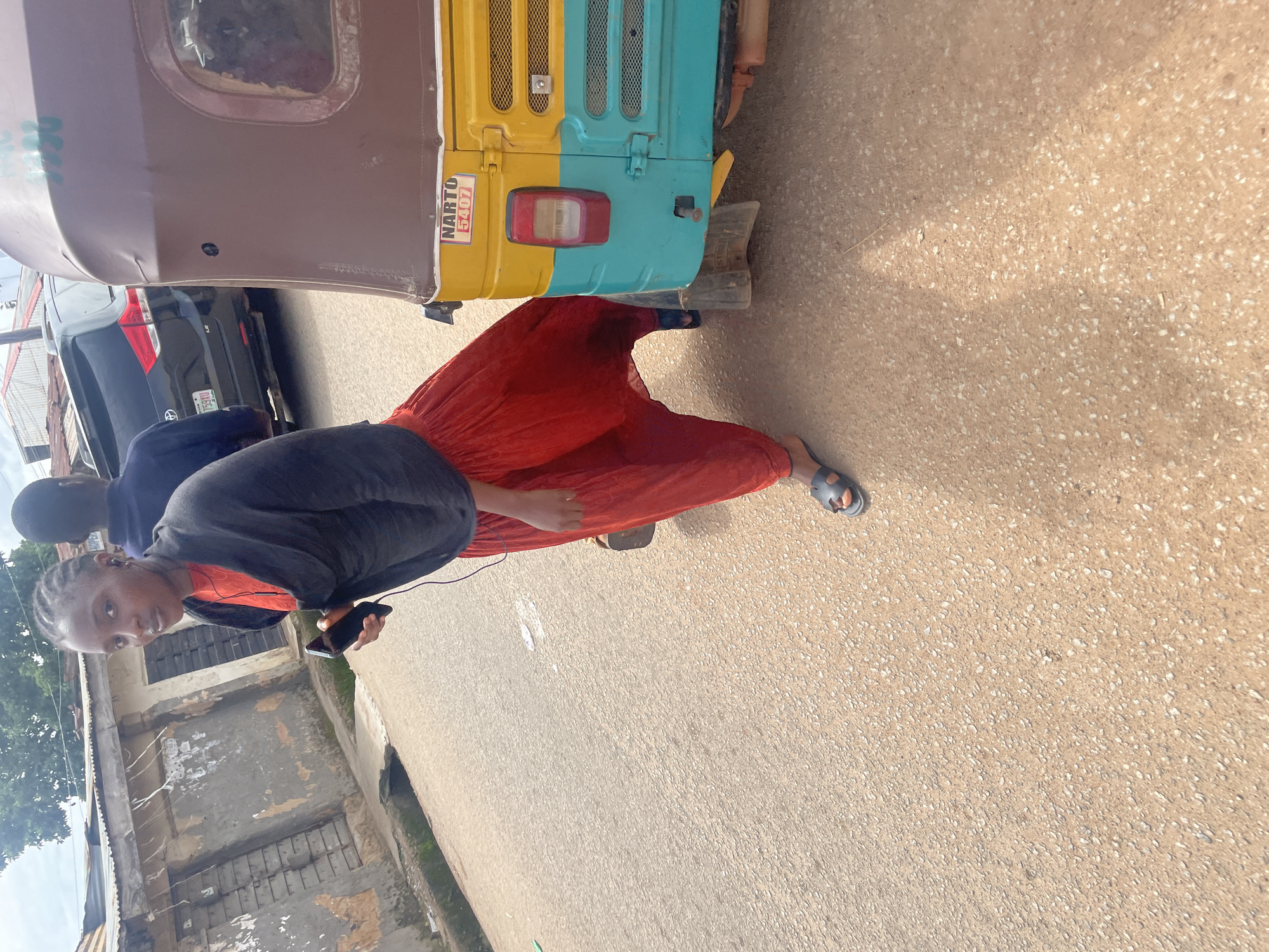 In Gura Top, Nigeria, a person stands on a dirt road next to a yellow and teal auto-rickshaw. They are wearing a red outfit and a black shawl, and are holding a smartphone in their hand. The auto-rickshaw has "NARTO 5407" written on its side. In the background, a black SUV is parked, and there are some buildings with weathered walls. The sky is overcast.