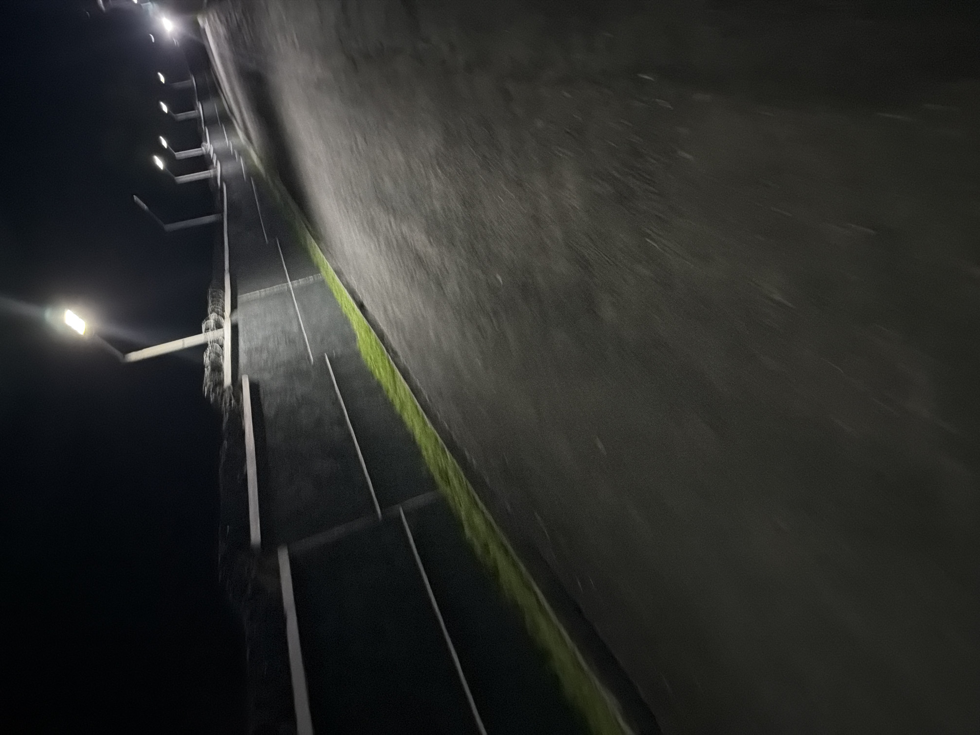 This image captures a dimly lit scene at night, likely in Bukuru, Nigeria, featuring a paved road or pathway that slopes gently upwards and to the right. The road surface is dark and appears somewhat textured. Along the left side of the road, a low, dark-colored wall runs parallel, with two thin white horizontal lines accentuating its surface. A strip of vibrant green grass or vegetation runs along the base of this wall.
Beyond the wall, the environment is darker, but several tall, slender lampposts with illuminated rectangular lights are visible, spaced at intervals along the upper edge of the wall. These lights cast a glow on the surrounding area. Further in the background, there are hints of more lampposts and possibly distant structures or vehicles, indicated by faint lights. The overall impression is one of an outdoor public space, possibly a walkway or street, illuminated by artificial lighting during nighttime hours.