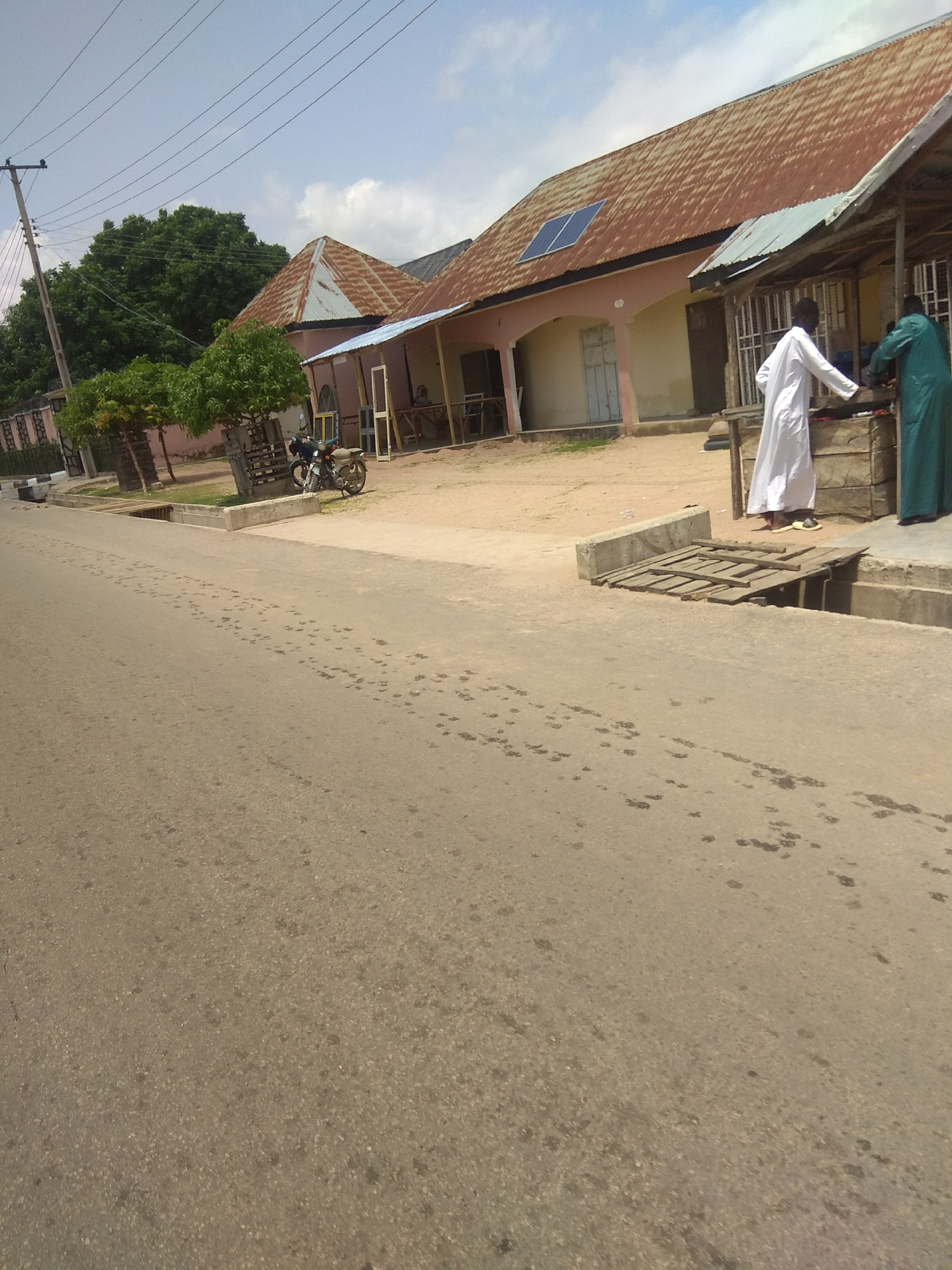 This is a daytime, medium-angle, outdoor photograph taken in Jalingo, Nigeria. The scene depicts a street with a road in the foreground, leading towards a building complex in the background. The building has a rusty, corrugated iron roof and a pale pink facade. A solar panel is visible on the roof. In front of the building, there is a covered wooden stall or shop. Two individuals are present; one person, wearing a white robe, is standing near the stall, and another person, wearing a green robe, is behind the stall, seemingly engaged in an activity. To the left of the building, there are trees and a motorcycle parked on the unpaved ground. A utility pole with wires is also visible on the left side of the image. The street has a drainage ditch with a wooden cover in the foreground. The sky is partly cloudy.