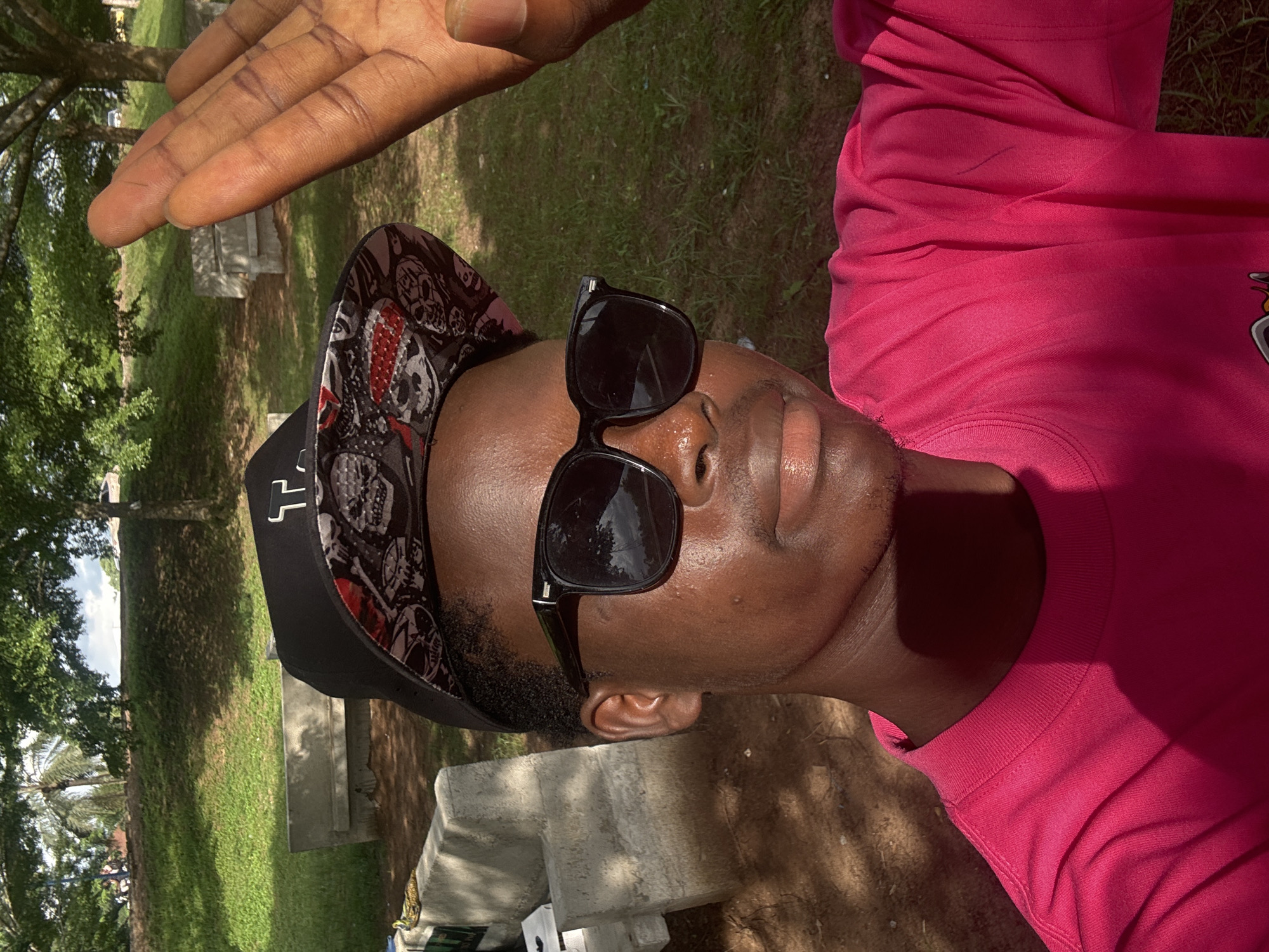 The image is a low-angle shot of a person with dark skin wearing a pink shirt, a black cap with a skull pattern, and black sunglasses. The person's hand is raised, casting a shadow on their face. The background shows a grassy area with trees and concrete structures, possibly in Ihiagwa, Nigeria.