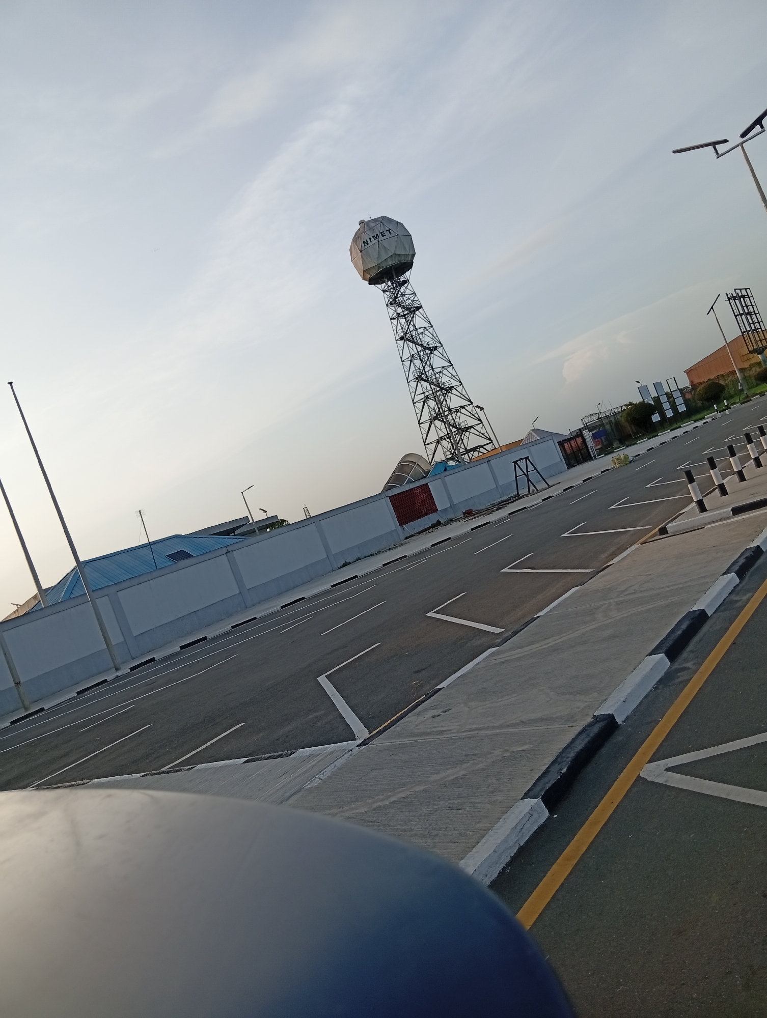 The image depicts a scene in Omagwa, Nigeria, featuring the Nigerian Meteorological Agency (NiMet) weather radar. A tall, steel lattice tower supports a large, spherical, white radome bearing the "NIMET" inscription. The structure is situated on a paved parking lot with parking spot markings and black-and-white painted bollards. A long, white concrete wall with black accents runs along the back of the parking area, in front of what appears to be an industrial or institutional facility with blue roofing. Several utility poles are also present. The sky is pale blue with light clouds. The bottom portion of the frame shows the hood of a vehicle in the foreground.