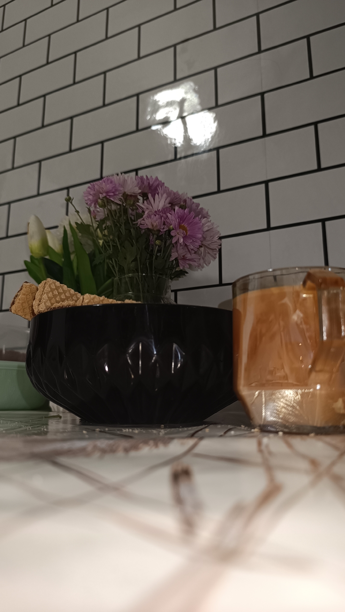 A close-up, low-angle shot captures a table setting against a white tiled wall with black grout. In the foreground, a glass filled with an amber-colored liquid and ice cubes is positioned to the right. To the left of the glass, a black, faceted bowl holds a bouquet of purple chrysanthemums and white tulips, with several wafer cookies resting on the rim of the bowl. The surface of the table is tiled with a white and grey pattern, with a blurry, abstract pattern overlaying it in the foreground. The lighting creates reflections on the wall and the bowl. The image appears to be taken indoors, possibly in a kitchen or dining area. There are no people or animals visible in the image. The location is not identifiable as Chaikovsky, Russia, based on the visual evidence.