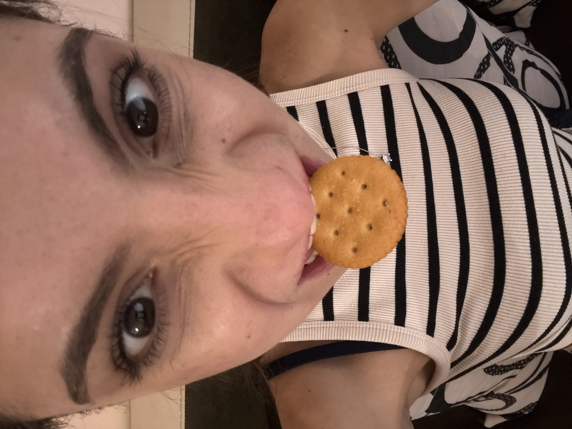 A woman is shown from a close, angled perspective, eating a round, tan cracker. The woman has dark eyes, thick eyebrows, and long eyelashes. She wears a white tank top with black vertical stripes. A thin, silver necklace with a small, clear gemstone is visible. Part of a black-and-white patterned garment is visible in the background. The context of the scene is not specified.