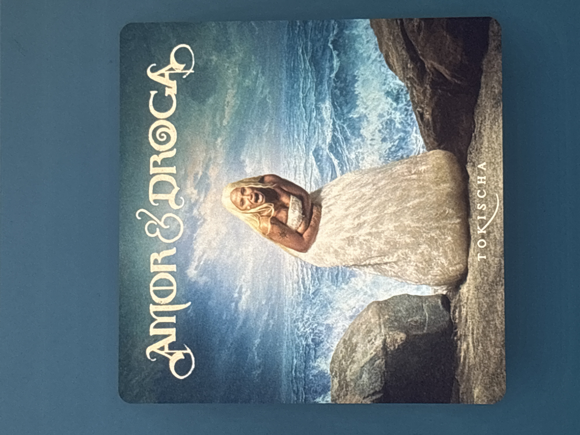 A vertical, eye-level shot depicts the cover of a music album. The cover art presents a woman in a white dress standing amidst rocks and a stylized ocean backdrop. The artist's name "TOKICHA" is vertically aligned on the right, and the title "AMOR & DROGA" is presented vertically on the left. The entire image is set against a solid blue background. The location is not discernible from the image.