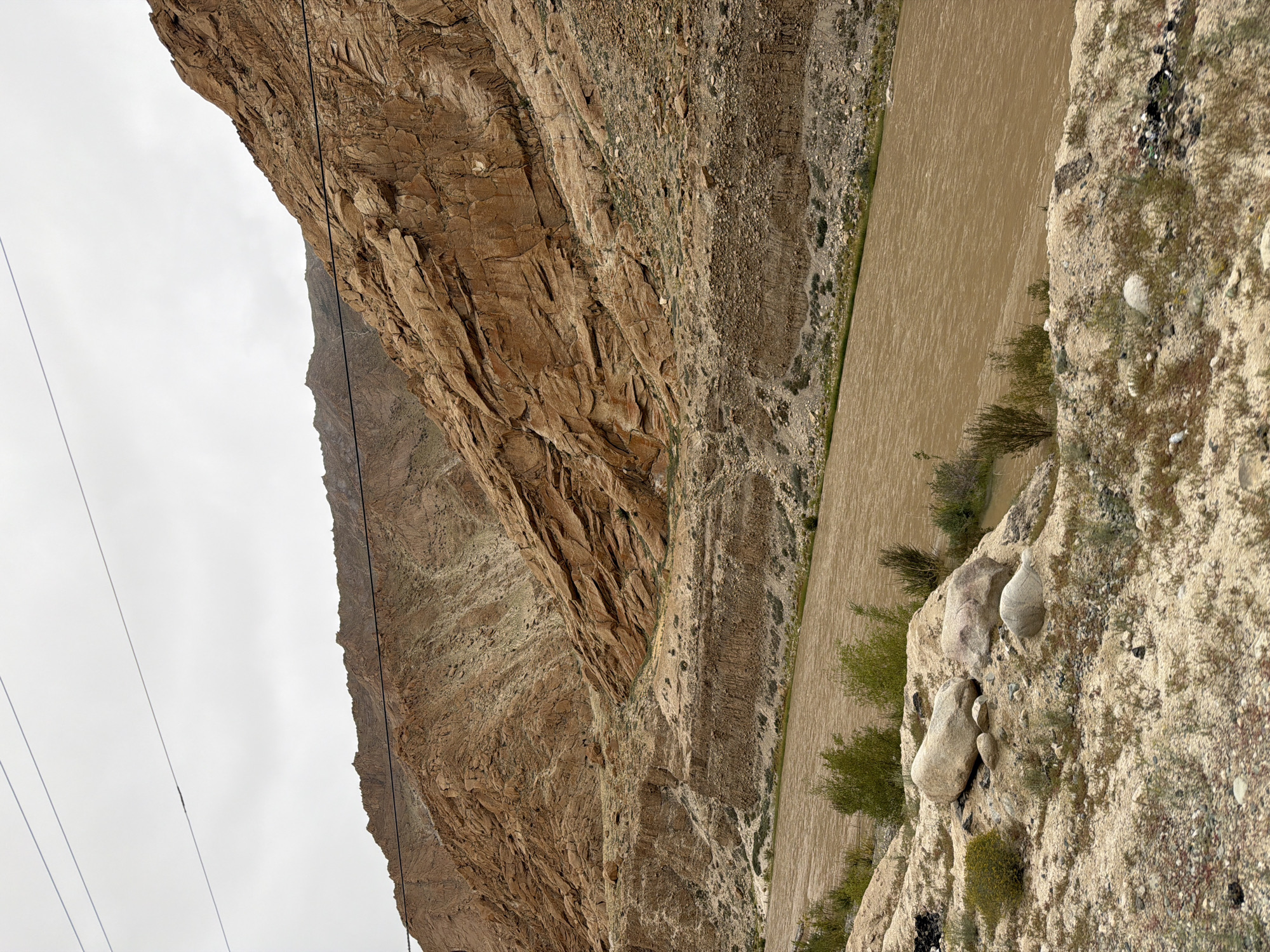 The image depicts a wide, muddy river flowing through a canyon with steep, rocky, and barren mountains on either side. The mountains are predominantly shades of brown and tan, with visible layers and striations in the rock formations. Sparse vegetation, consisting of low bushes and grasses, can be seen on the slopes and along the riverbanks. In the foreground, there is dry, sandy ground with a few scattered rocks and small plants. Overhead, utility lines are visible against a cloudy sky. The overall scene suggests a remote and rugged natural landscape. The location is identified as Hymia, India. There are no people visible in the image.