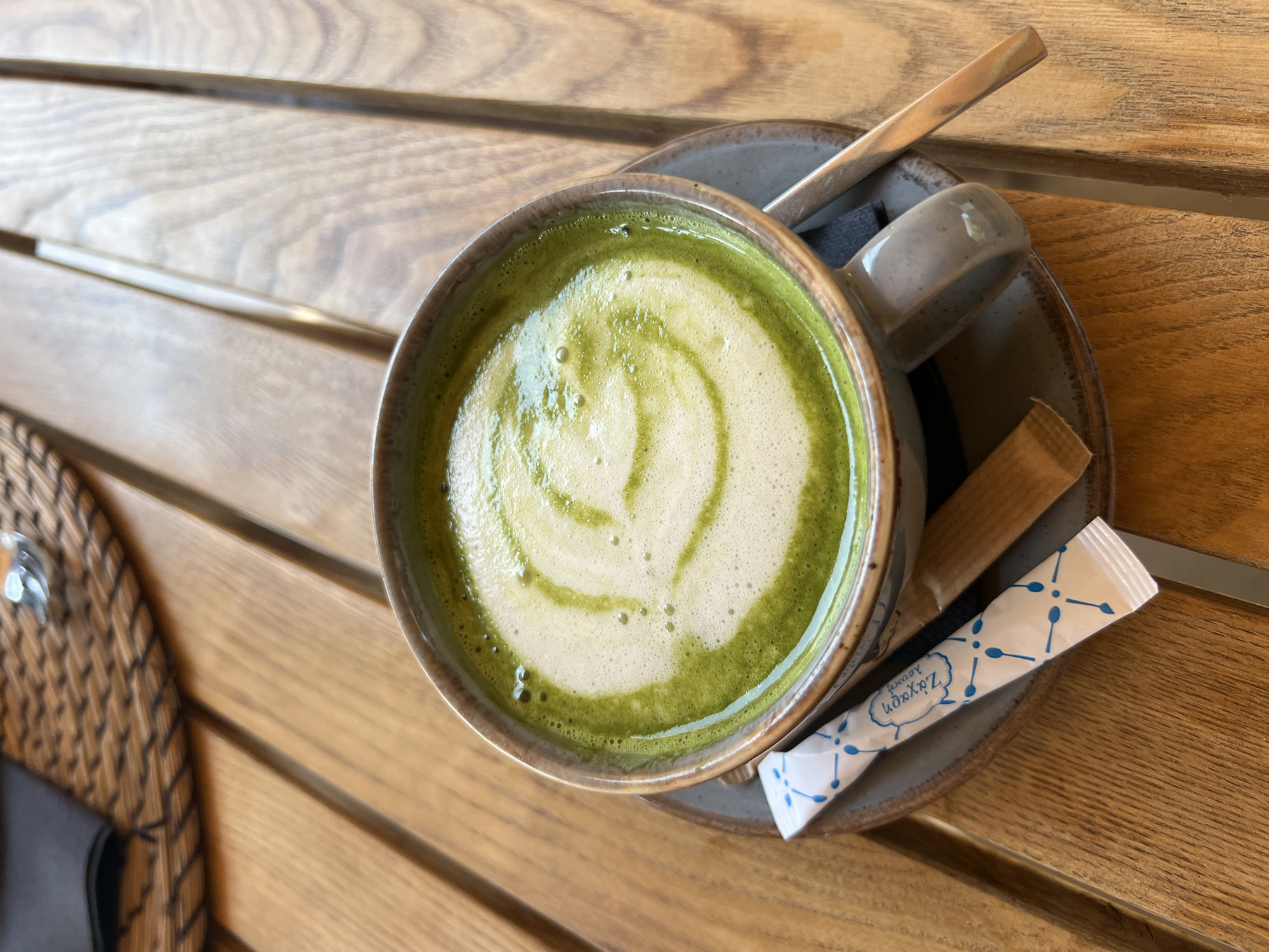 A close-up, overhead shot shows a steaming cup of matcha latte on a wooden table. The latte has a frothy, white top with a green swirl pattern. A small sugar packet with blue designs and a wooden stirrer are placed on the saucer next to the cup. A metallic spoon rests on the saucer, partially submerged in the liquid. The table is made of light brown wooden planks with visible grain. In the background, a woven placemat and a black napkin are visible. The scene appears to be set in a cafe in Naousa, Greece.
