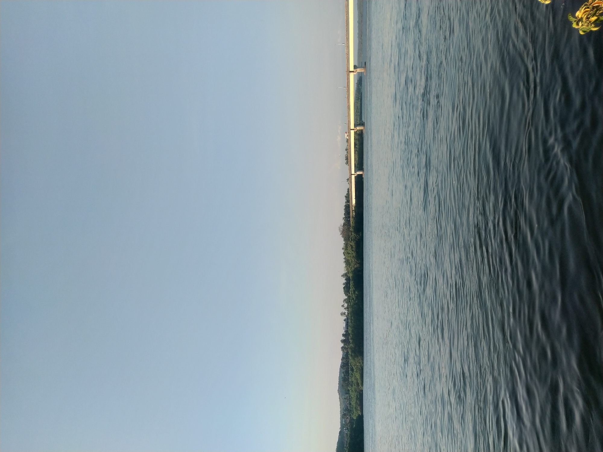 The image depicts a body of water, possibly a river or lake, on the right side. The water surface shows signs of ripples or waves. On the left is a clear sky. A strip of land with trees runs between the water and the sky. A bridge structure can be seen spanning the water in the distance. The image's location is Oxel, India.