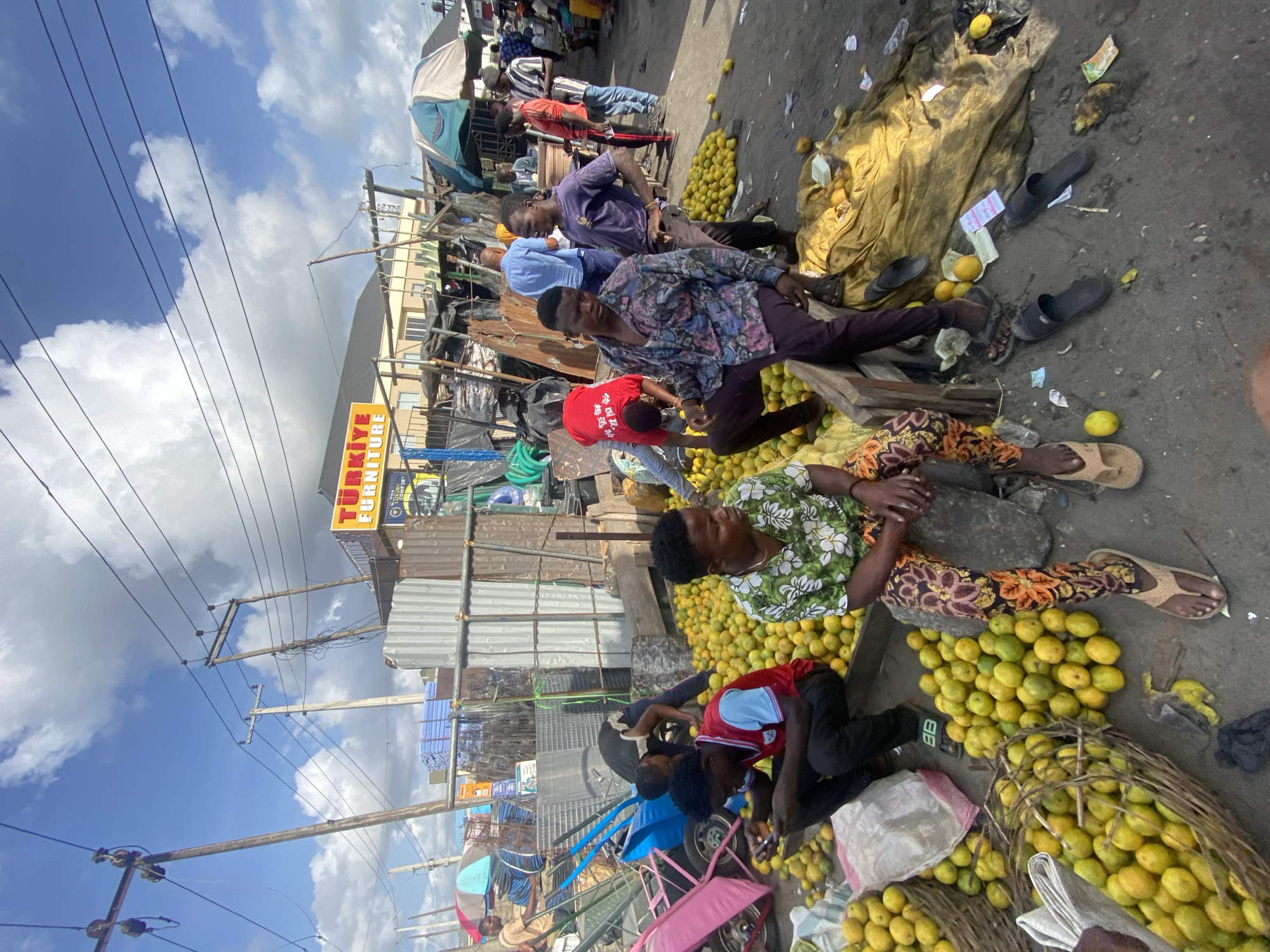 In an outdoor market scene in Lekki, Nigeria, several individuals are positioned amongst a large quantity of lemons. Some are seated directly on piles of lemons, while others are sorting or handling them. The street market setup includes various stalls with corrugated metal roofing and tarpaulin coverings. A sign for “TURKIYE FURNITURE” is visible above the market. Overhead, there are utility poles and wires against a backdrop of a cloudy sky. The ground surface is asphalt with scattered debris. Baskets are filled with lemons, suggesting they are the primary commodity being sold or traded.